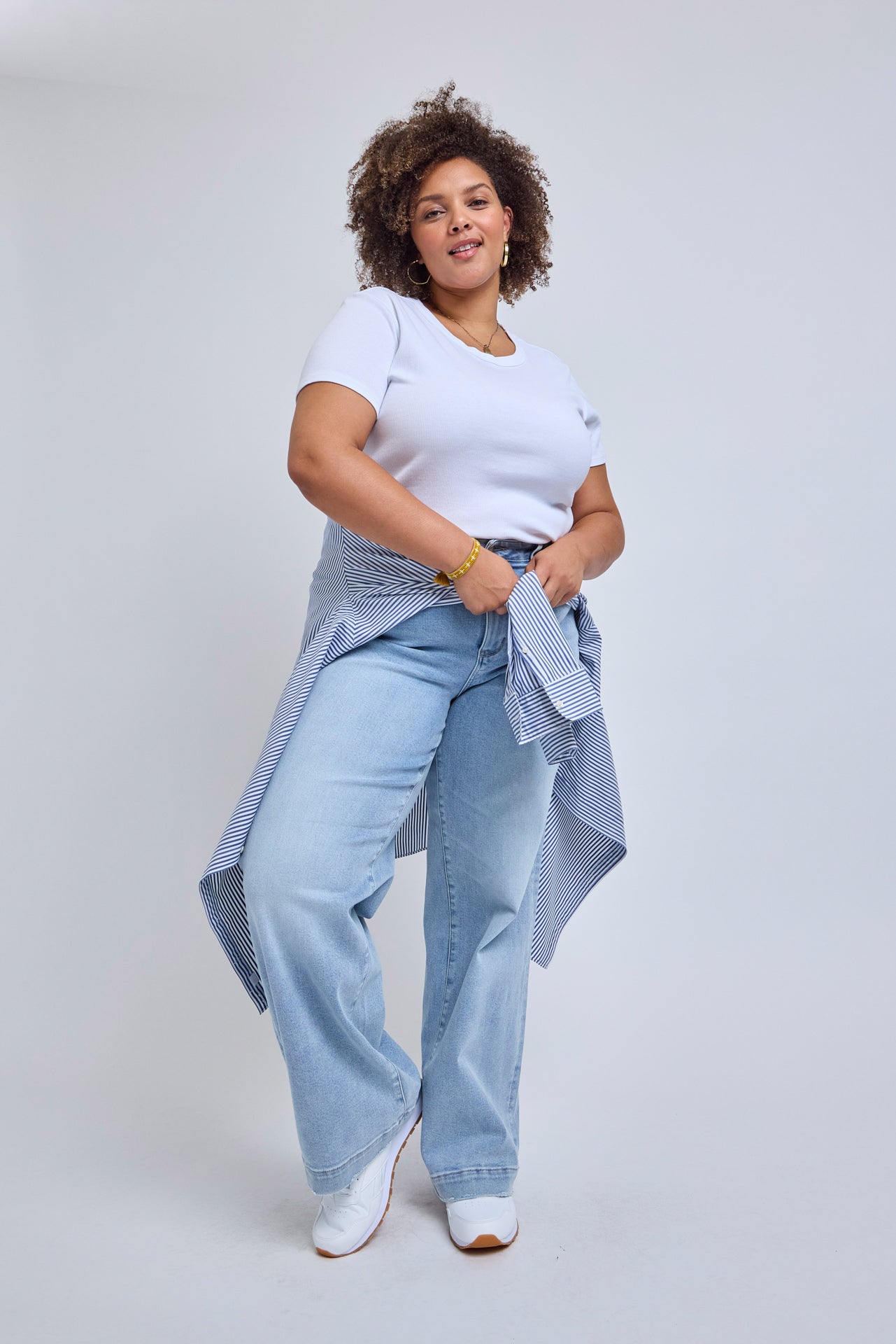 Woman wearing a white t-shirt, light blue jeans, and a plaid shirt over her shoulder on a plain background | model-size: 16