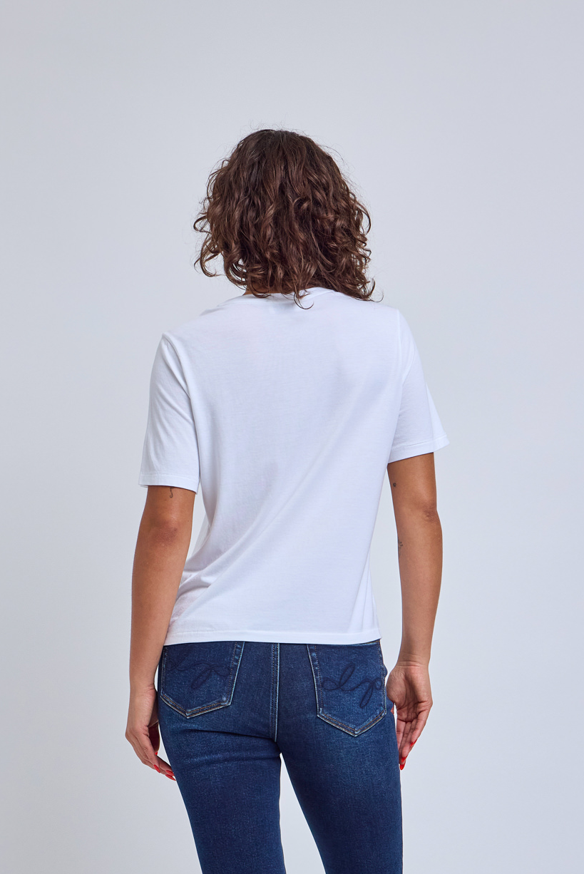 Back side picture of a woman wearing a white crew neck shirt and jeans against a gray wall