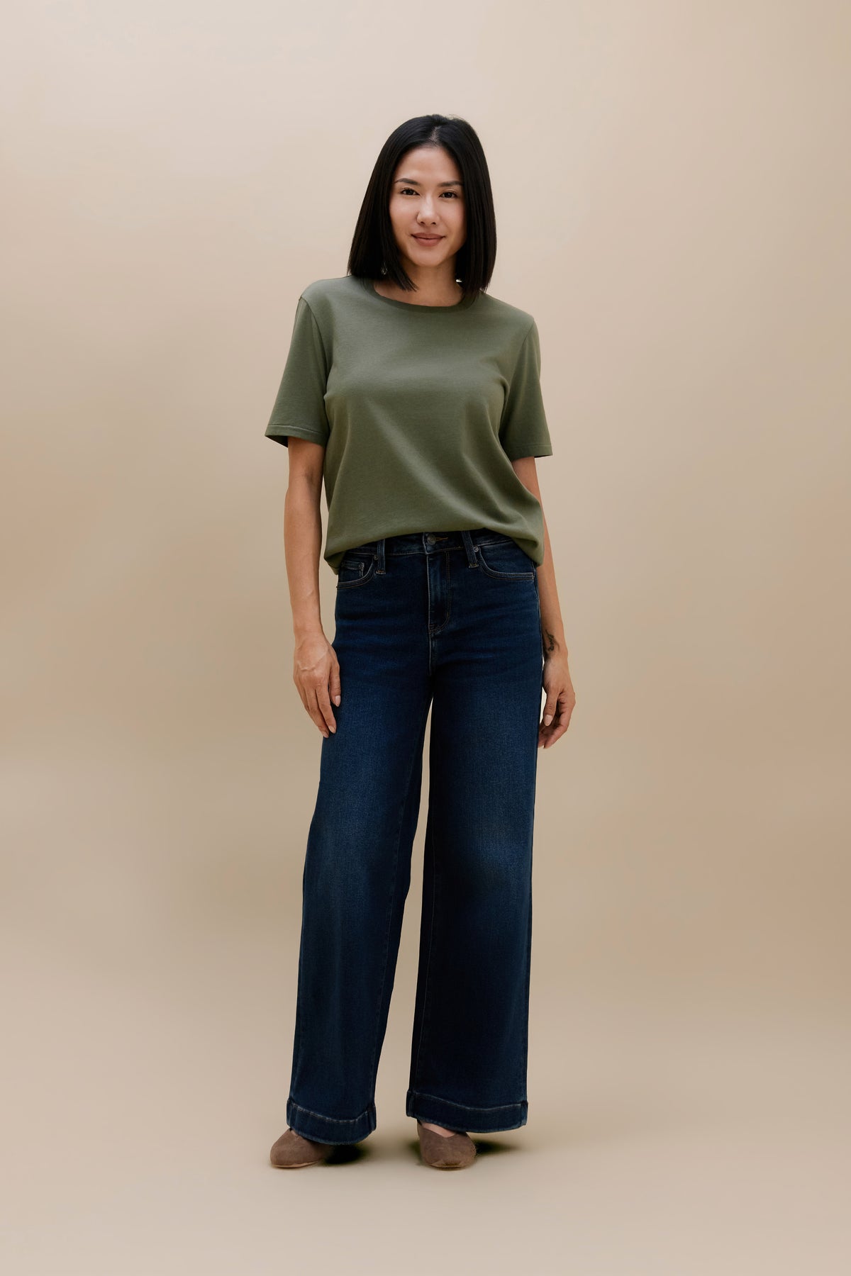 A woman modeling a short-sleeve olive green t-shirt and dark wide-leg denim jeans against a solid beige studio background.