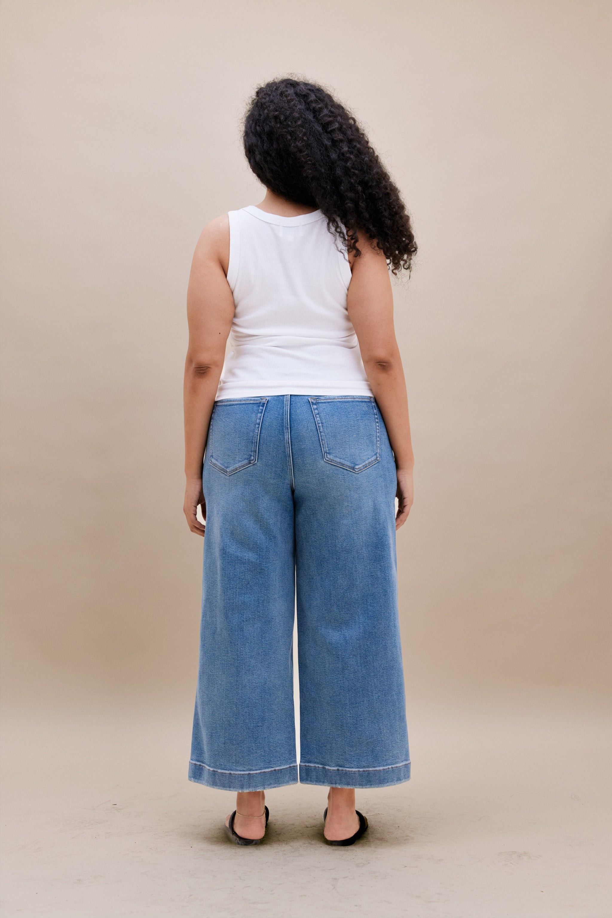 Backside of a woman wearing a white tank top with wide leg cropped jeans with a beige background