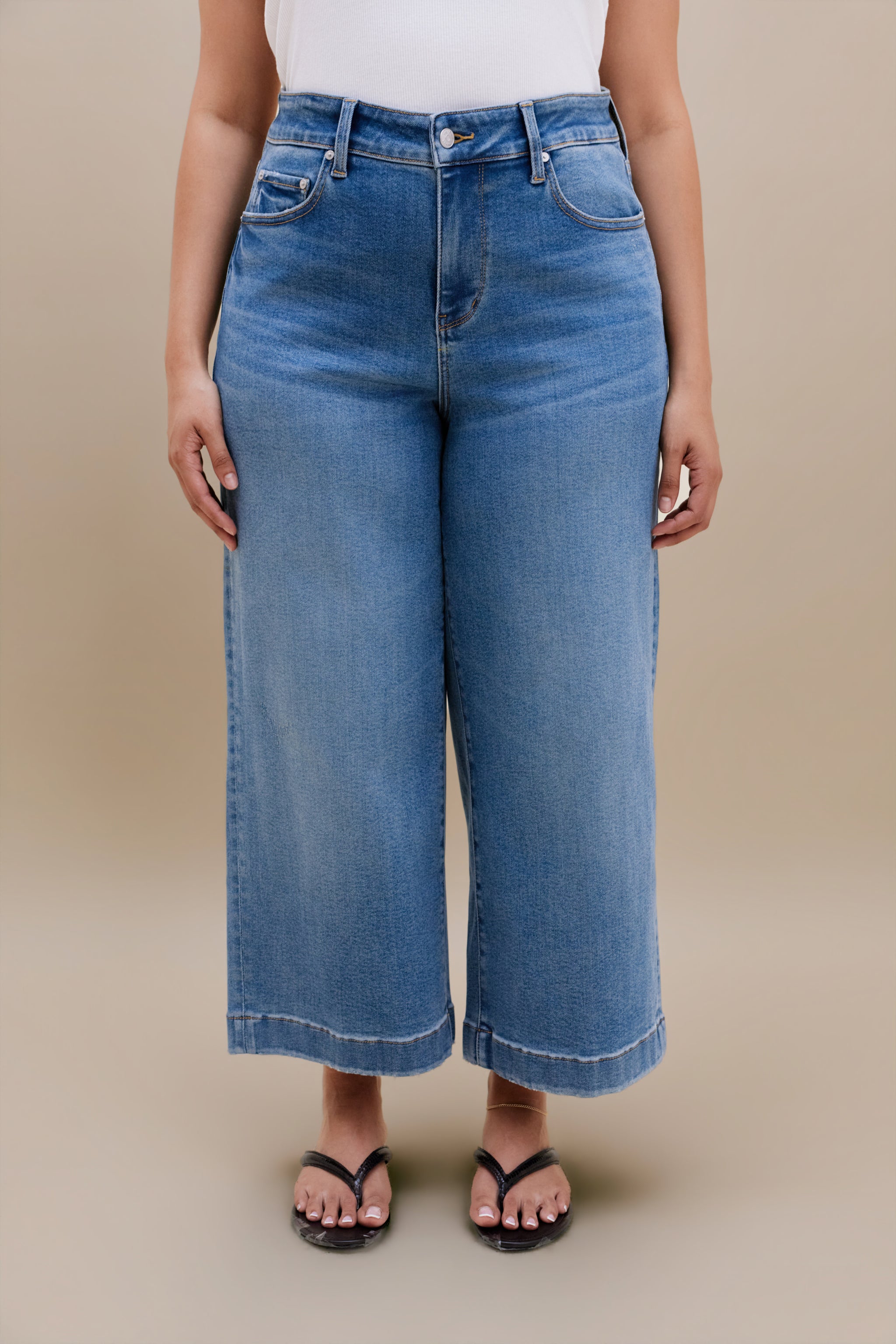 Bottom half of a woman wearing a white tank top with wide leg cropped jeans with a beige background