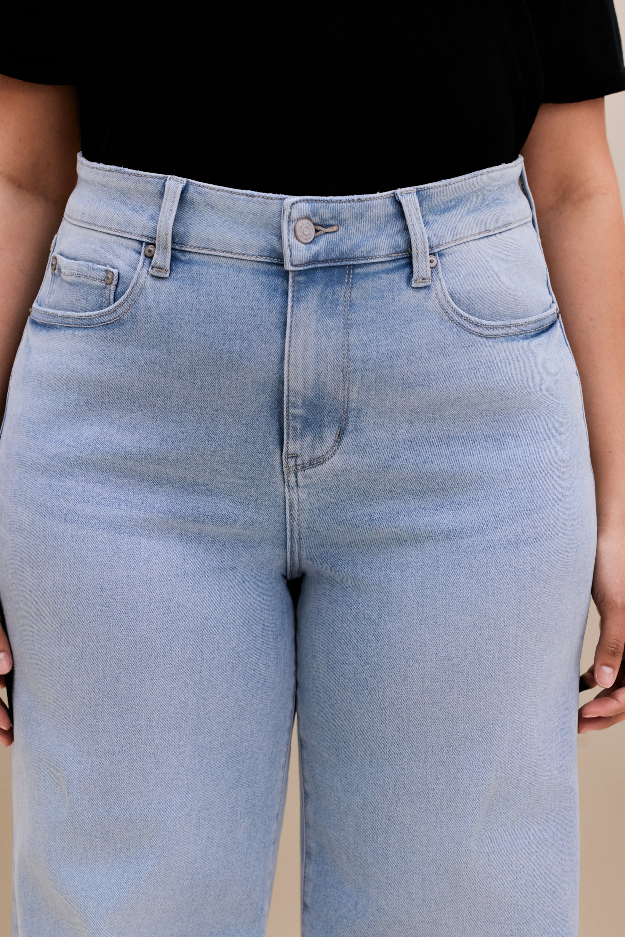 Close up highlighting the waist of a woman wearing a black v-neck t-shirt and light colored wide leg jeans with a beige background
