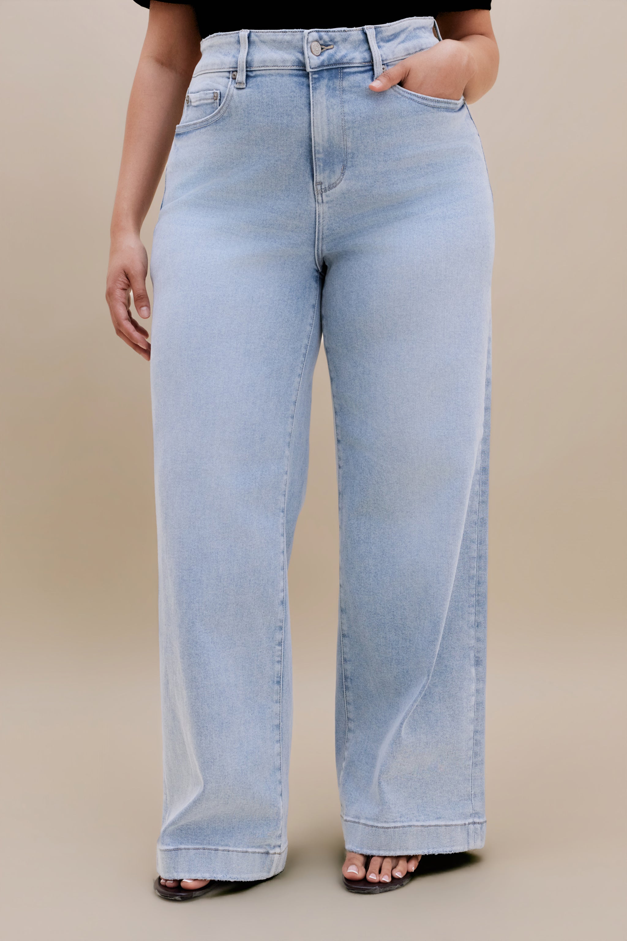 Bottom half or a woman wearing a black v-neck t-shirt and light colored wide leg jeans with a beige background