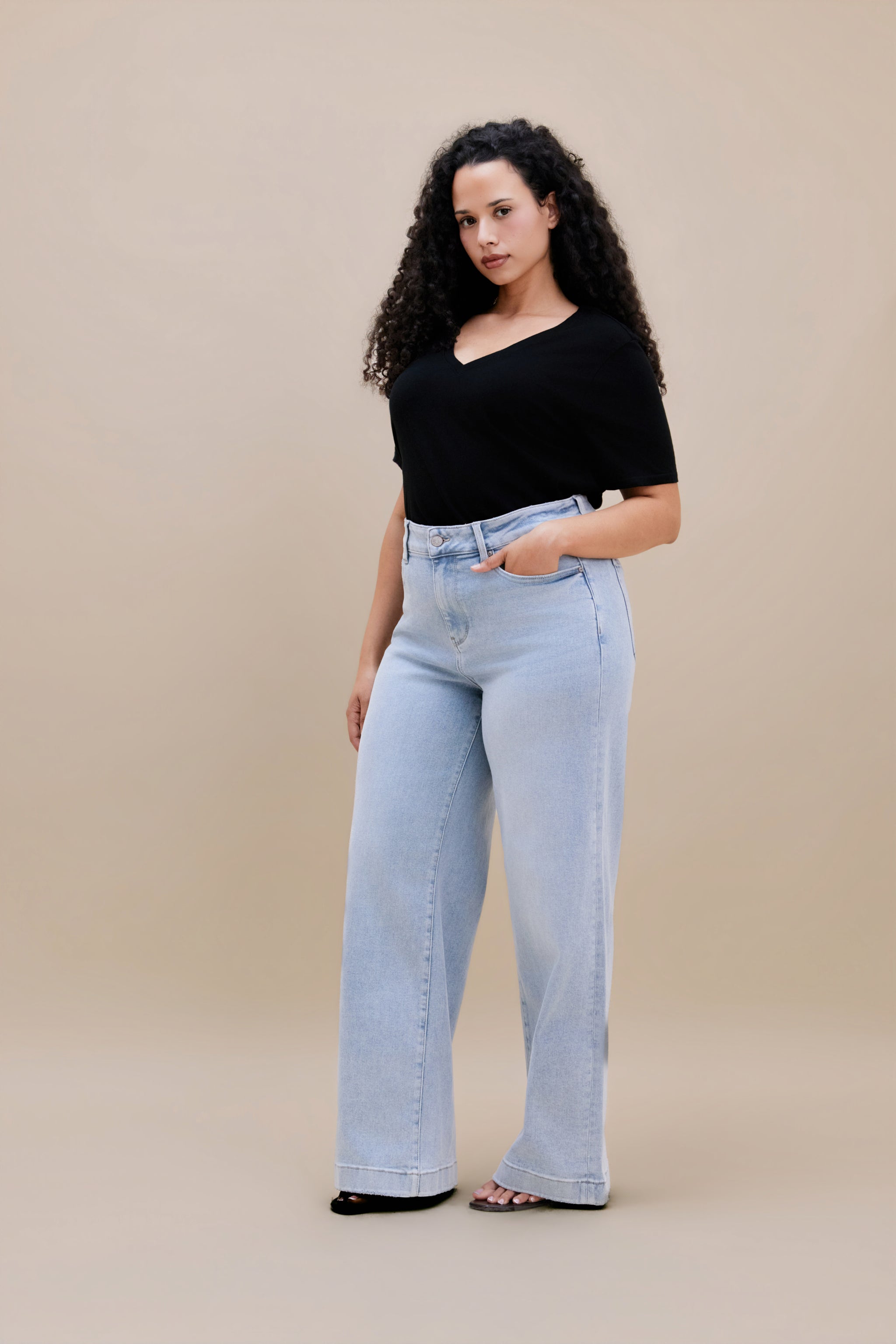 Woman wearing a black v-neck t-shirt and light colored wide leg jeans with a beige background