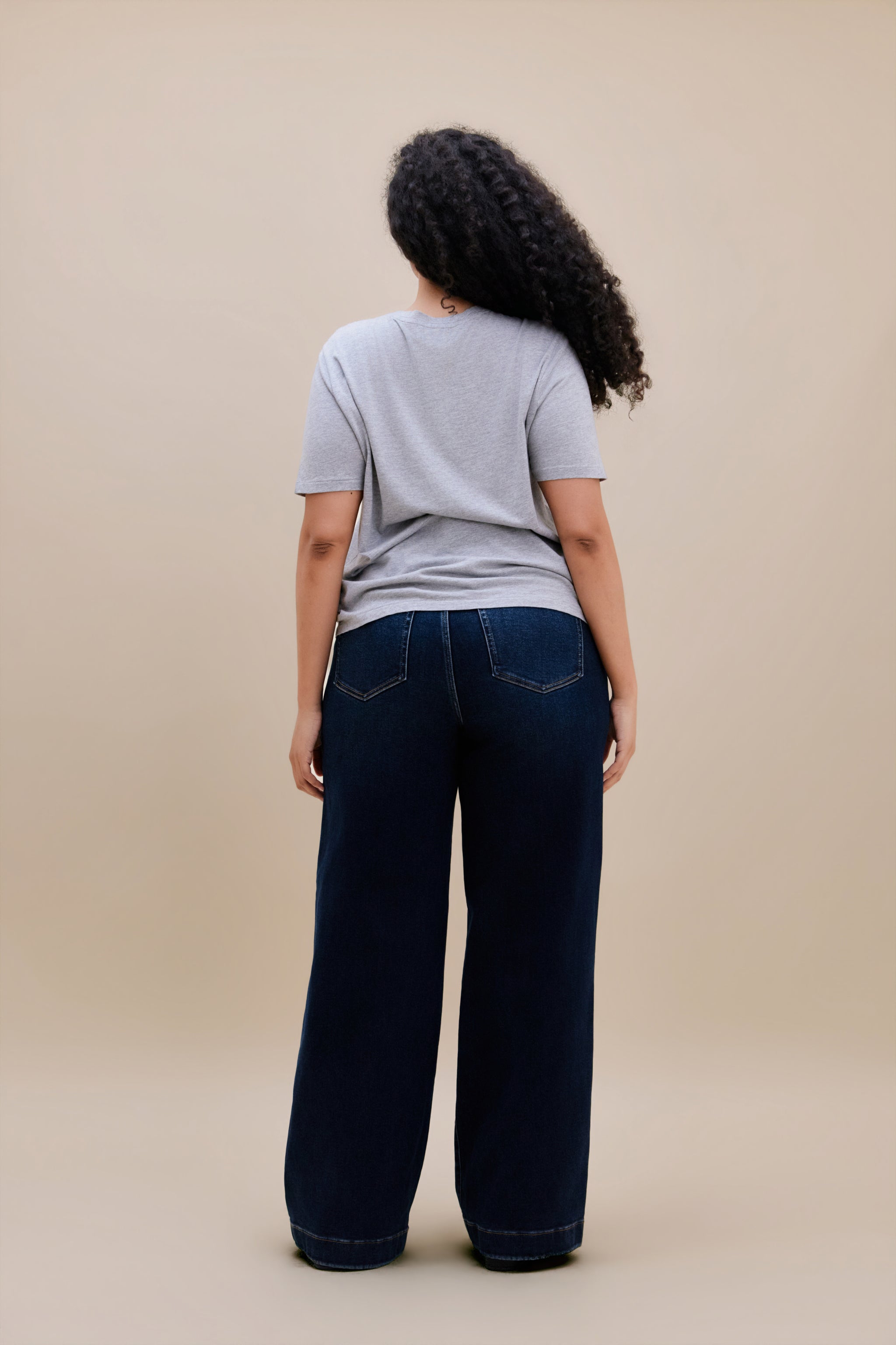 Woman wearing a gray t-shirt and dark wide leg jeans standing with a beige background