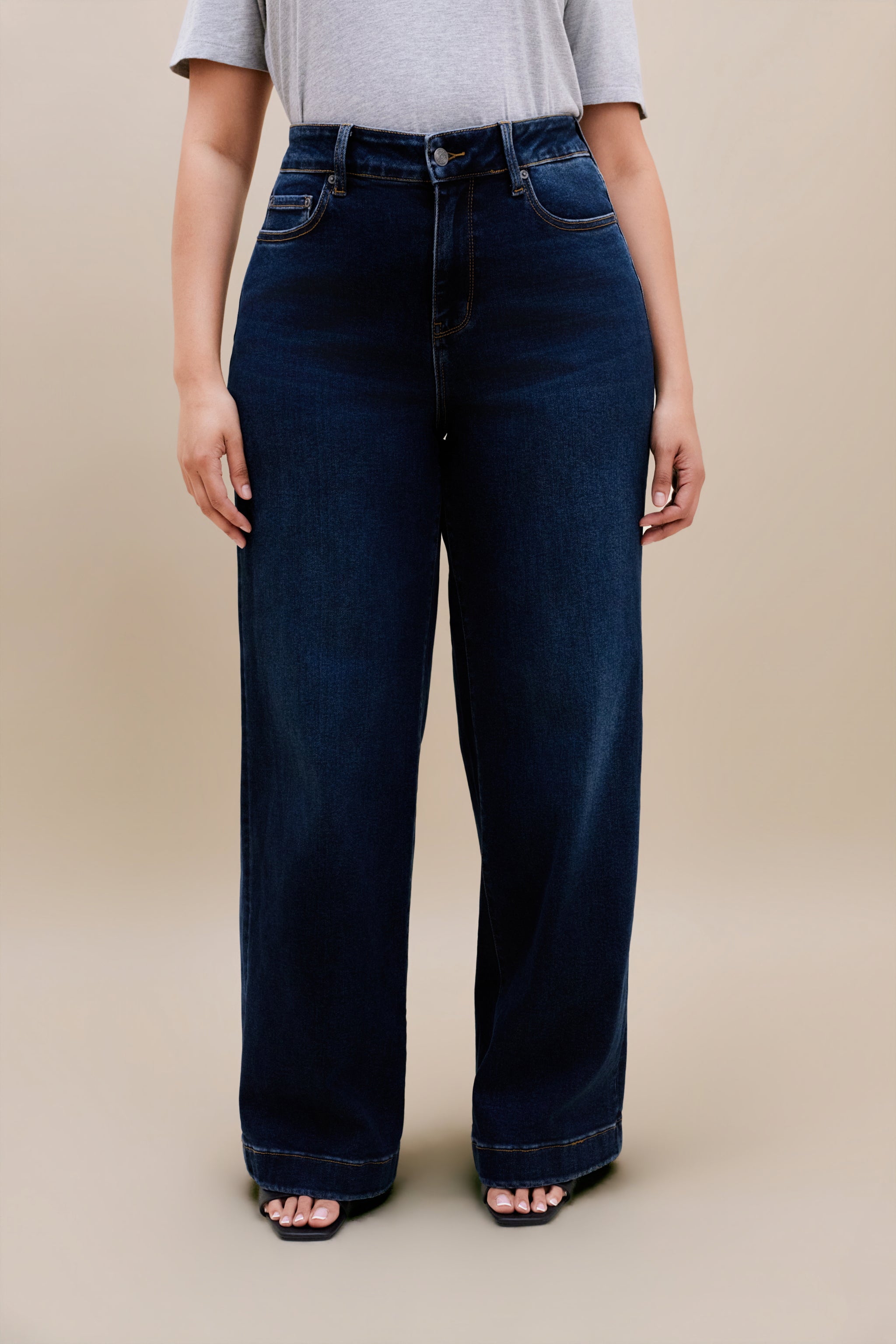 Woman wearing a gray t-shirt and dark wide leg jeans standing with a beige background