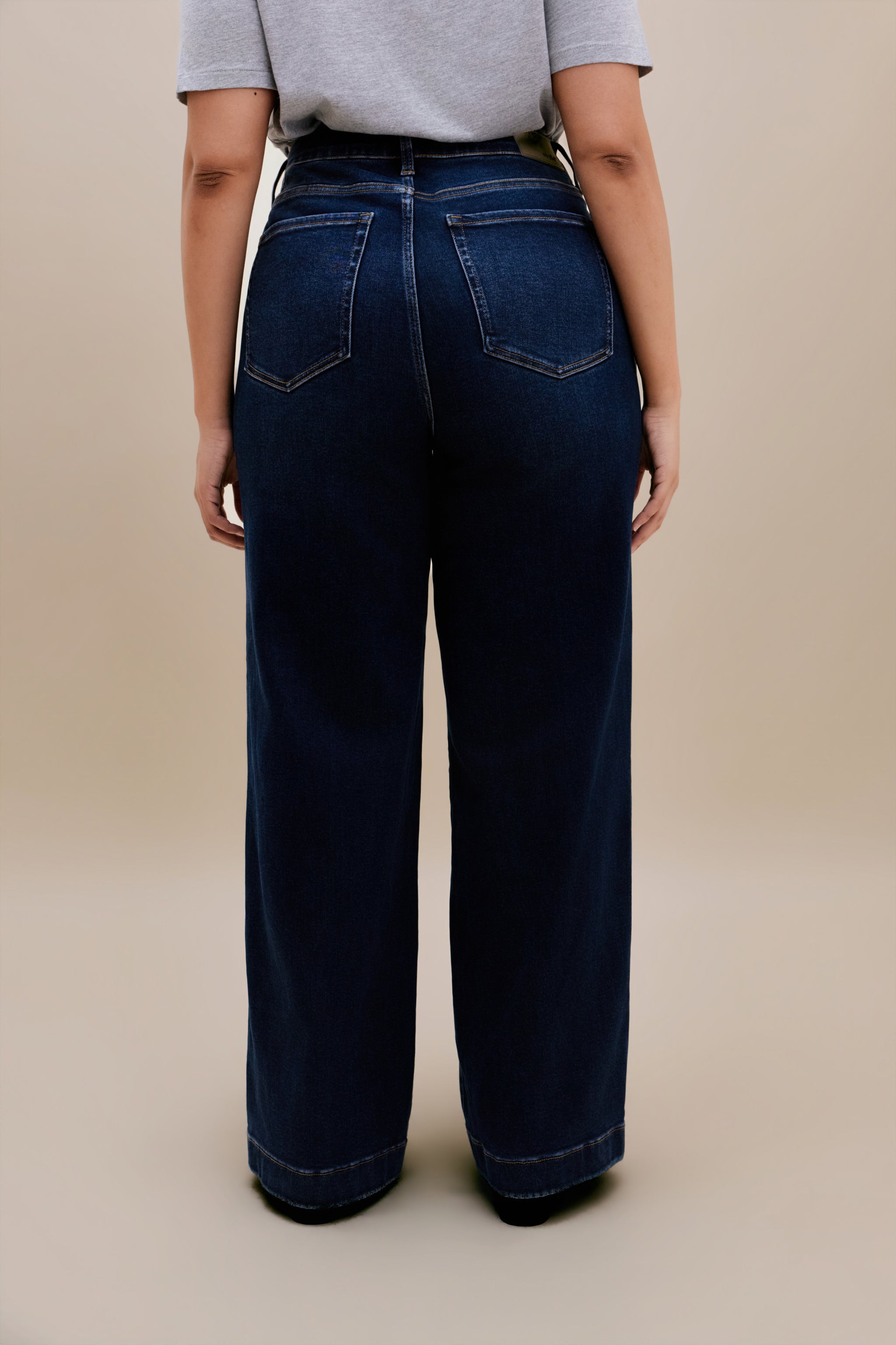 Woman wearing a gray t-shirt and dark wide leg jeans standing with a beige background