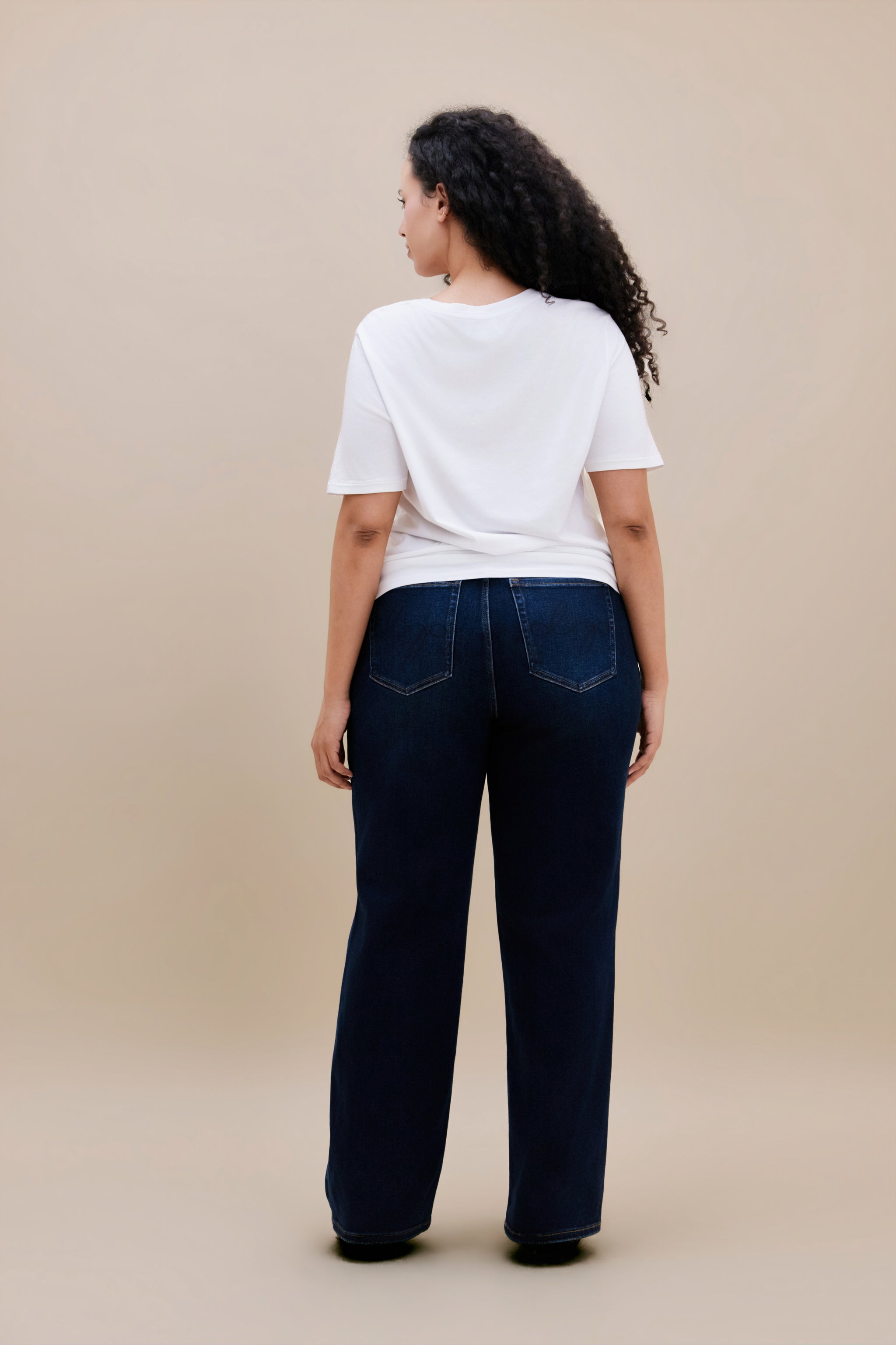 Woman wearing white t-shirt and dark straight leg jeans with a beige background