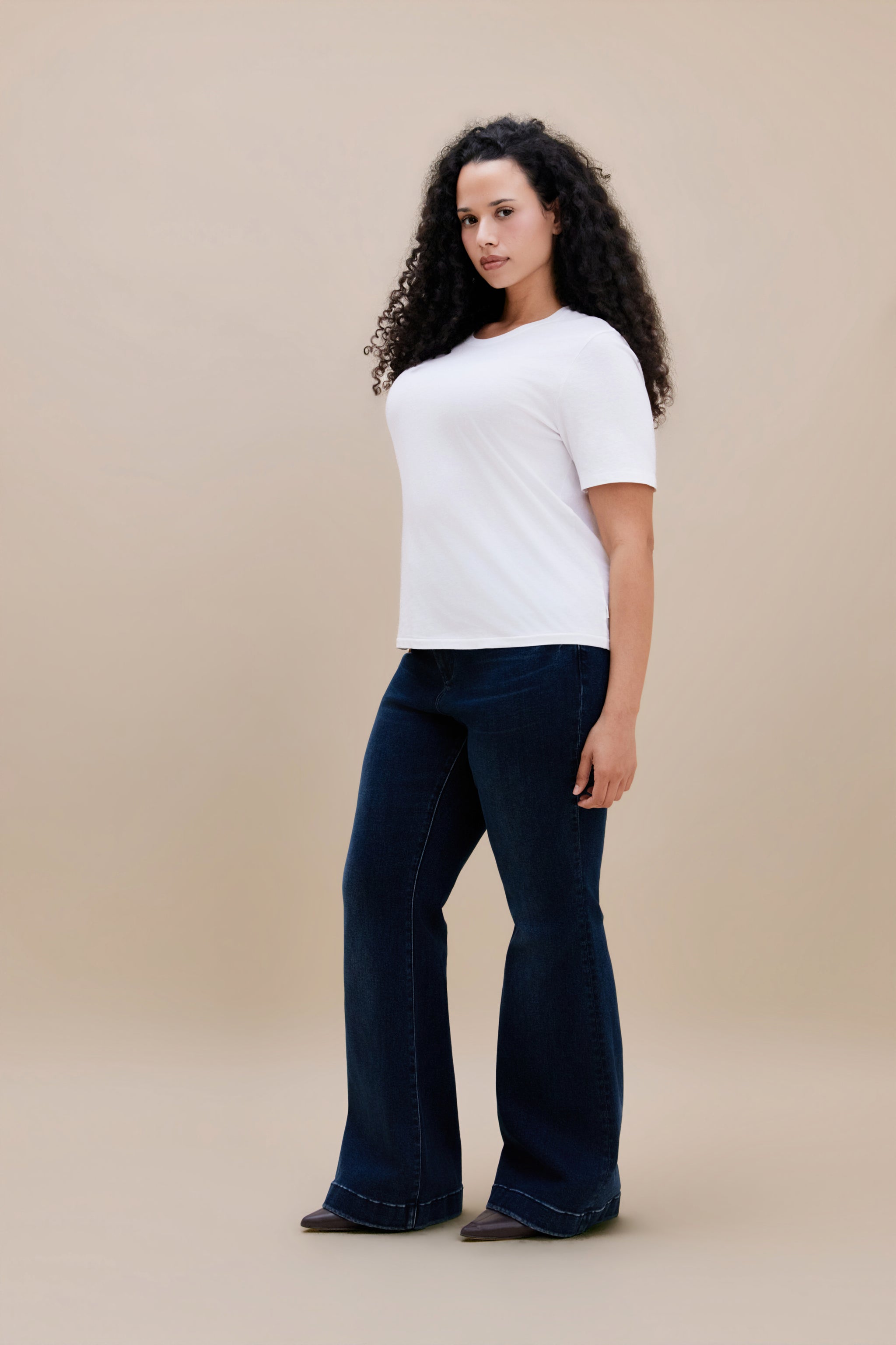 Woman wearing a white t-shirt and dark flare jeans with a beige background