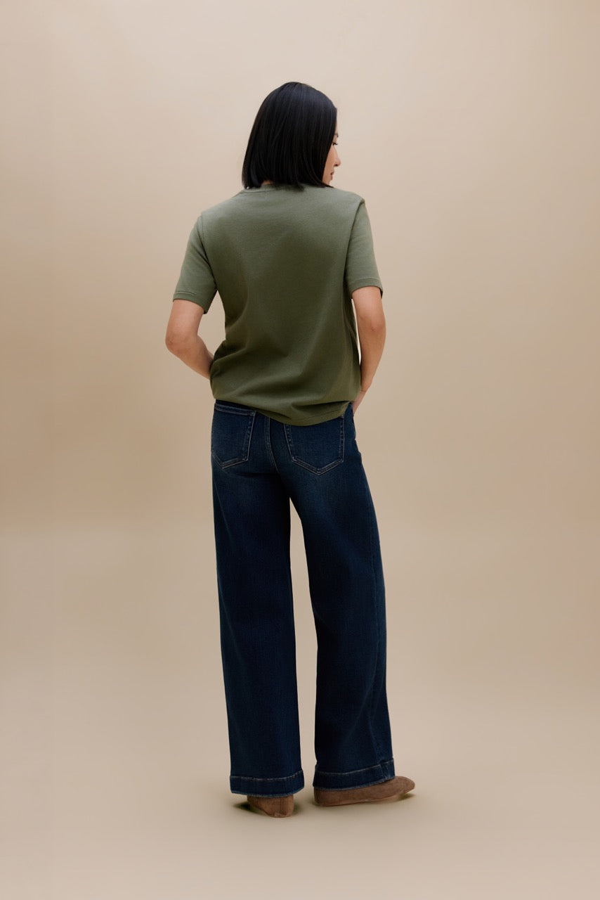 A model shown from the back wearing an olive green short-sleeve t-shirt and dark blue wide-leg jeans, standing against a solid beige studio background.