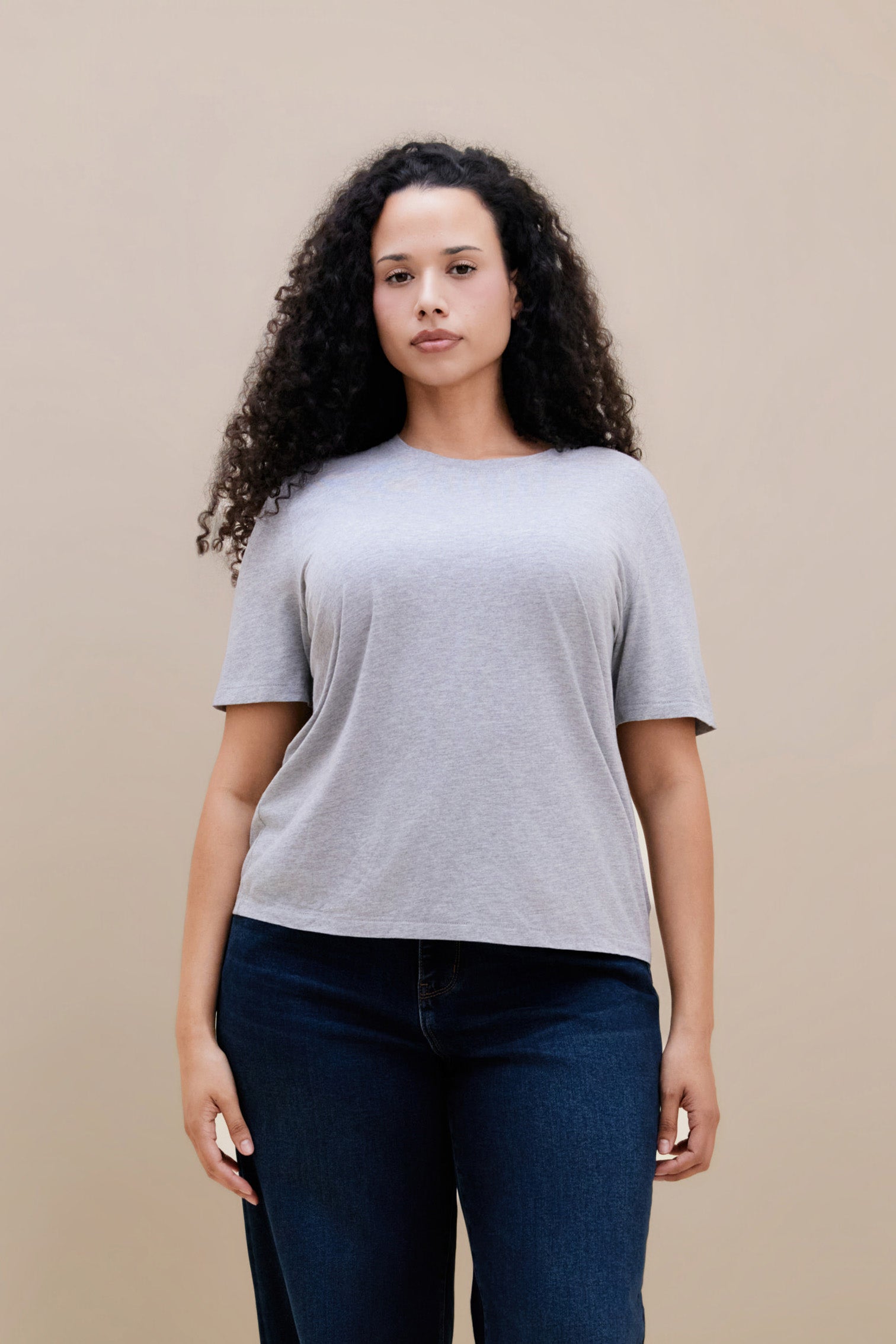 Woman wearing a light heather grey t-shirt & dark jeans against a beige background | model-size: 16