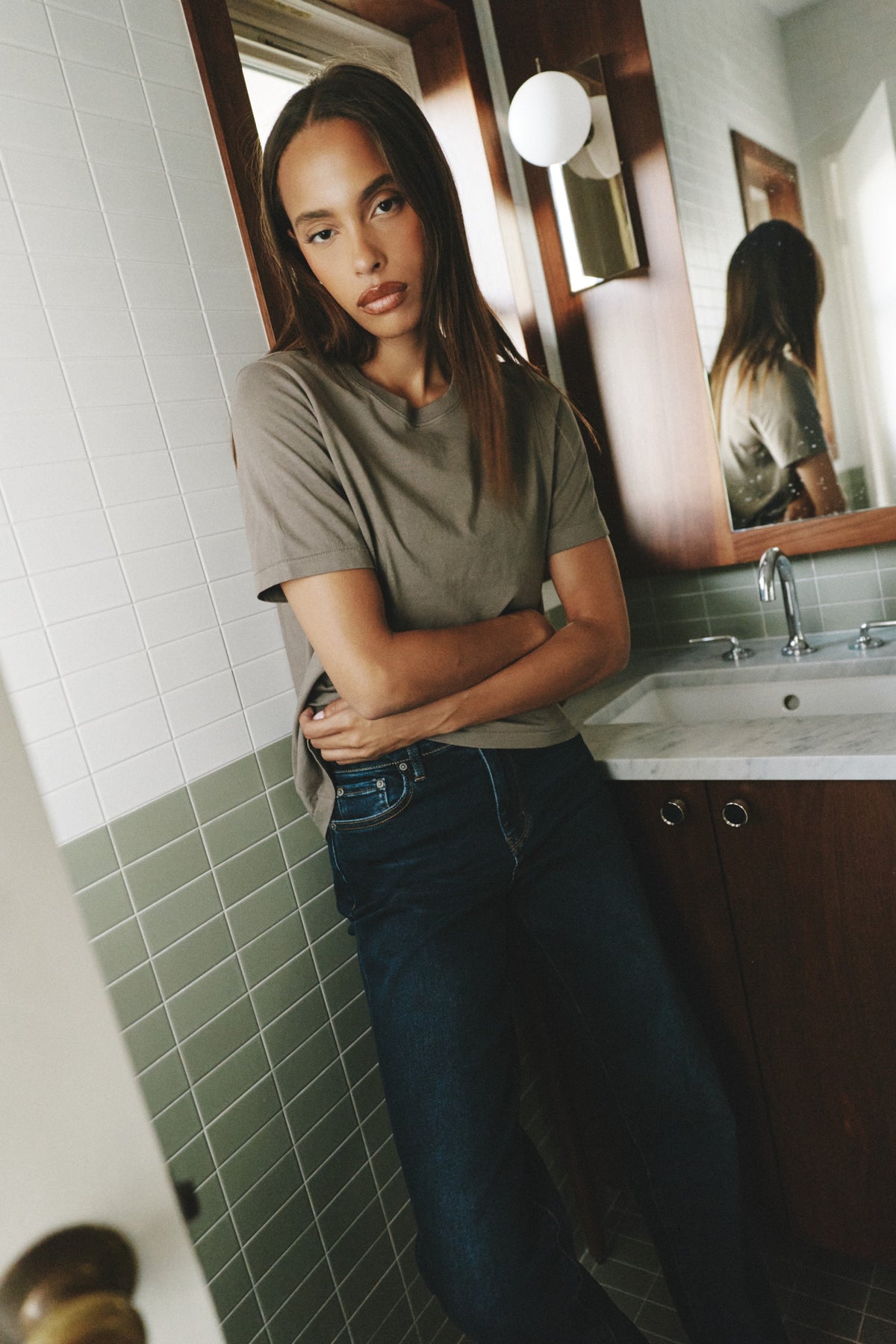 A woman with long dark hair stands in a tiled bathroom wearing a muted olive-gray short-sleeve t-shirt and dark blue jeans, with a sink and mirror visible behind her.