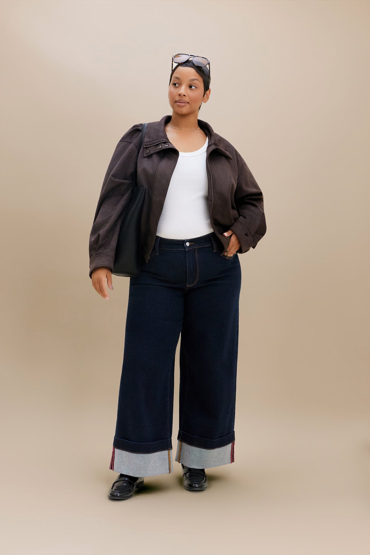 A woman modeling a dark brown cropped jacket, dark wide-leg jeans with rolled cuffs, a white tank top, and black shoes against a solid beige studio background.