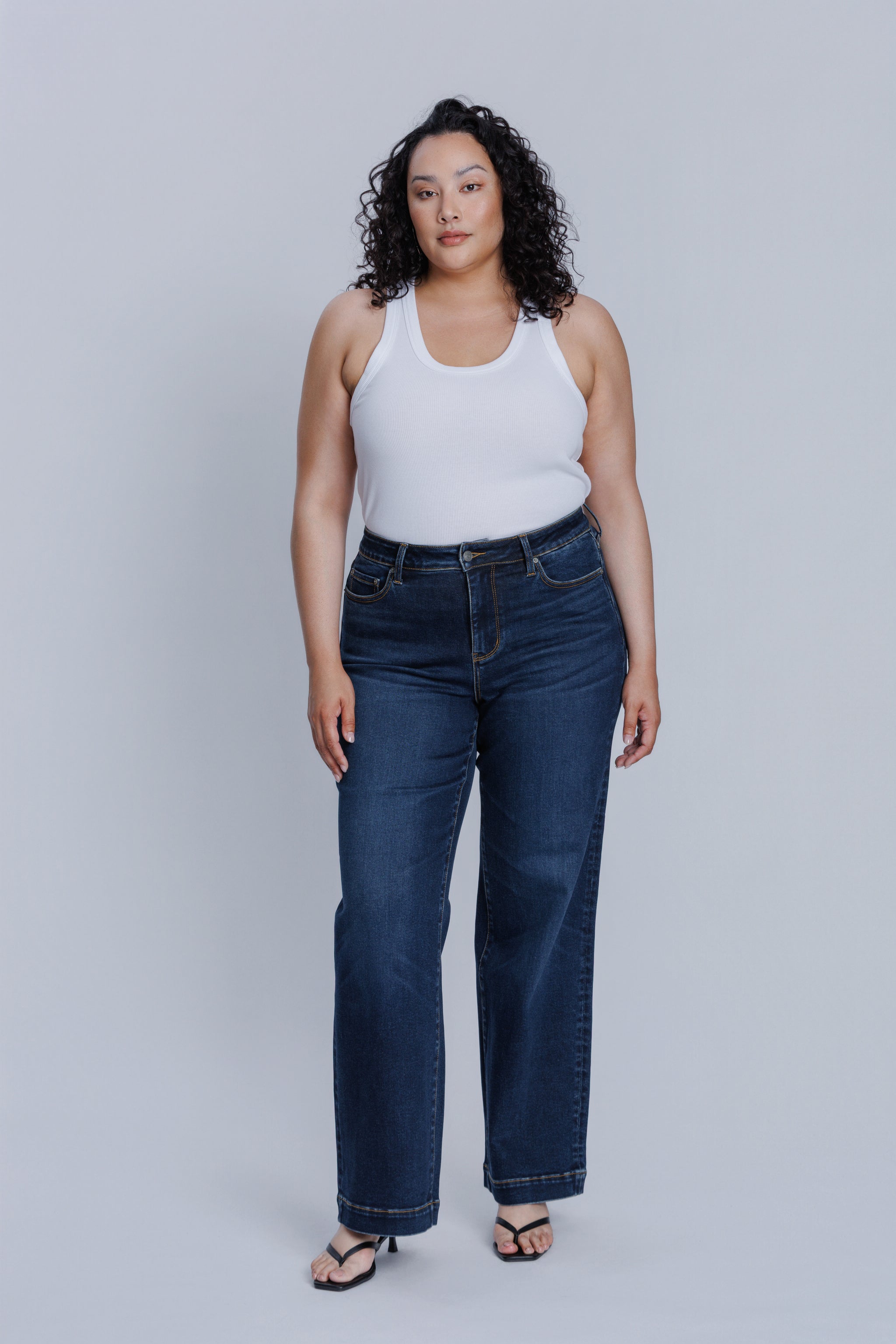 Woman wearing a DePart white tank top and dark blue jeans against a light gray background | model-size: 16