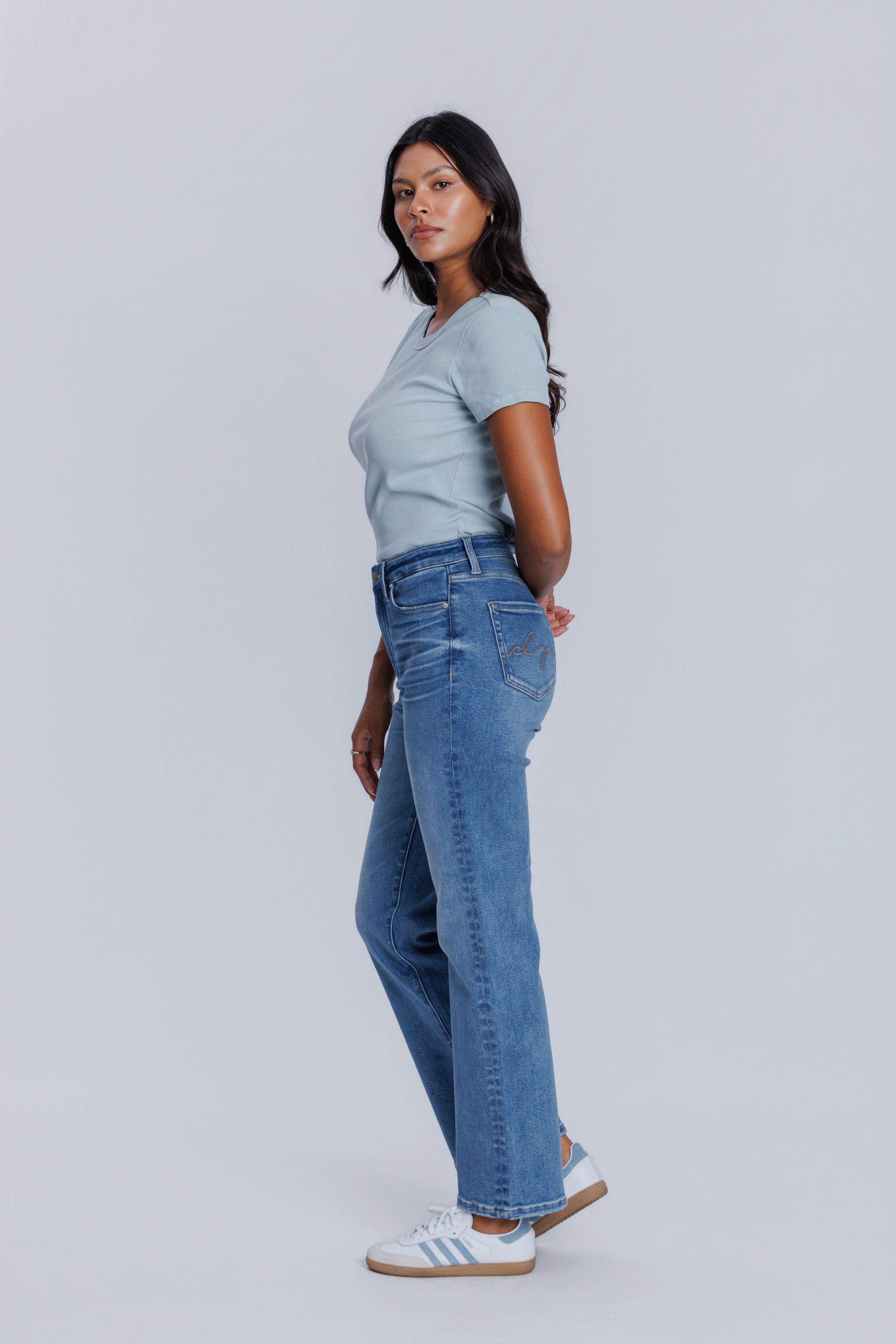 Woman standing sideways wearing a DePart light blue tee and blue jeans against a plain background