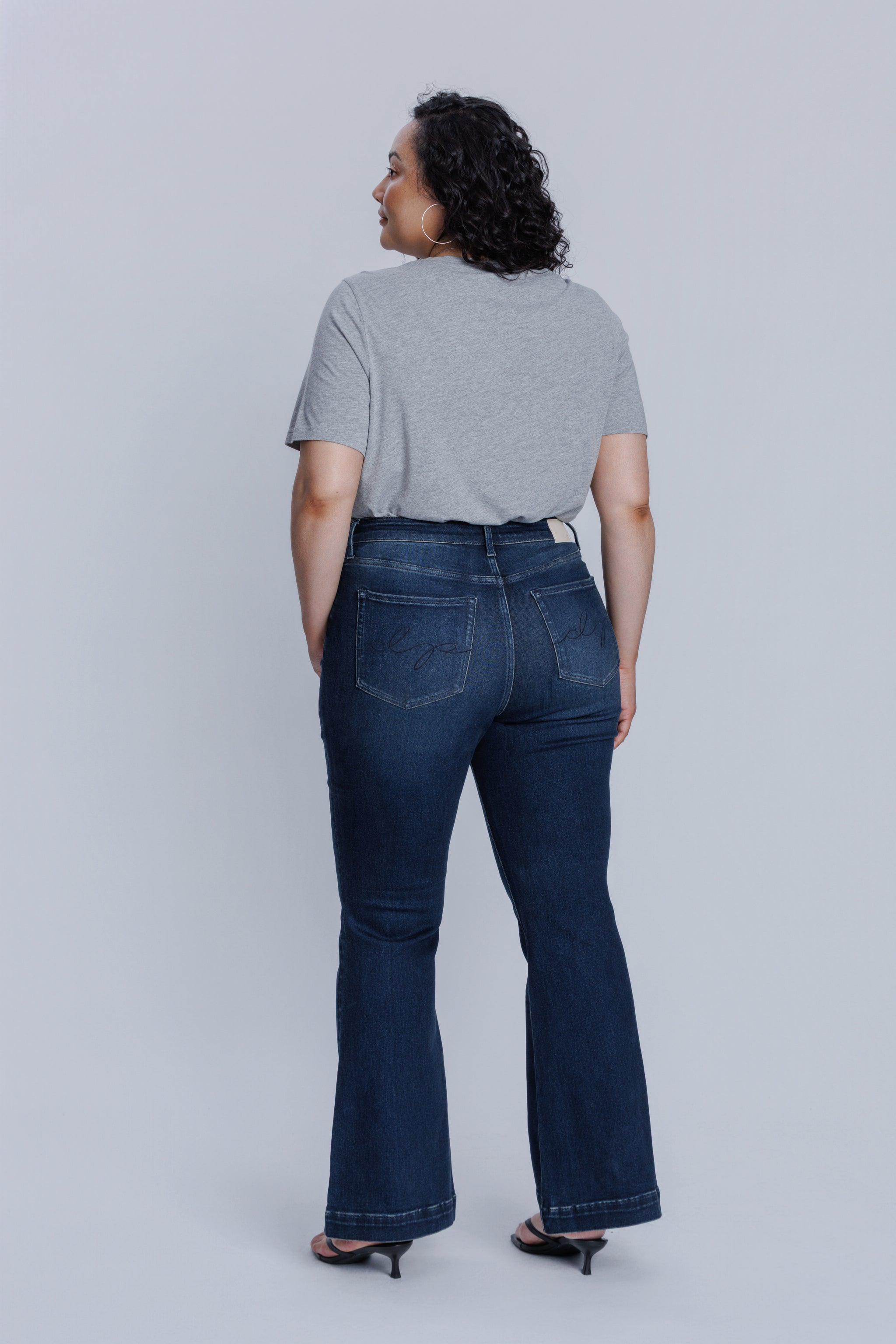 Woman wearing a DePart gray crew neck t-shirt and dark blue jeans against a light gray background