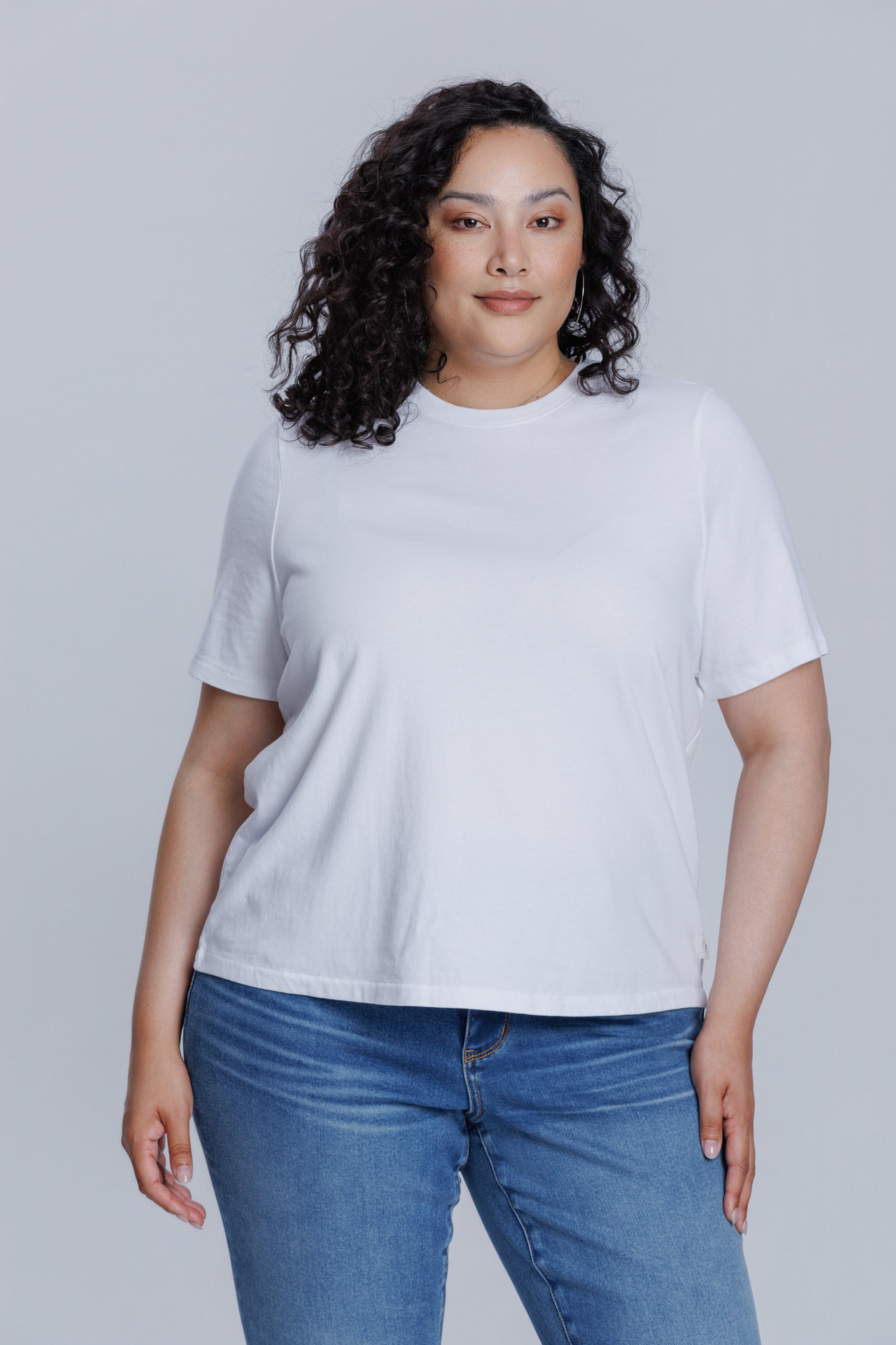 Woman wearing a DePart white t-shirt and blue jeans against a light gray background | model-size: 16