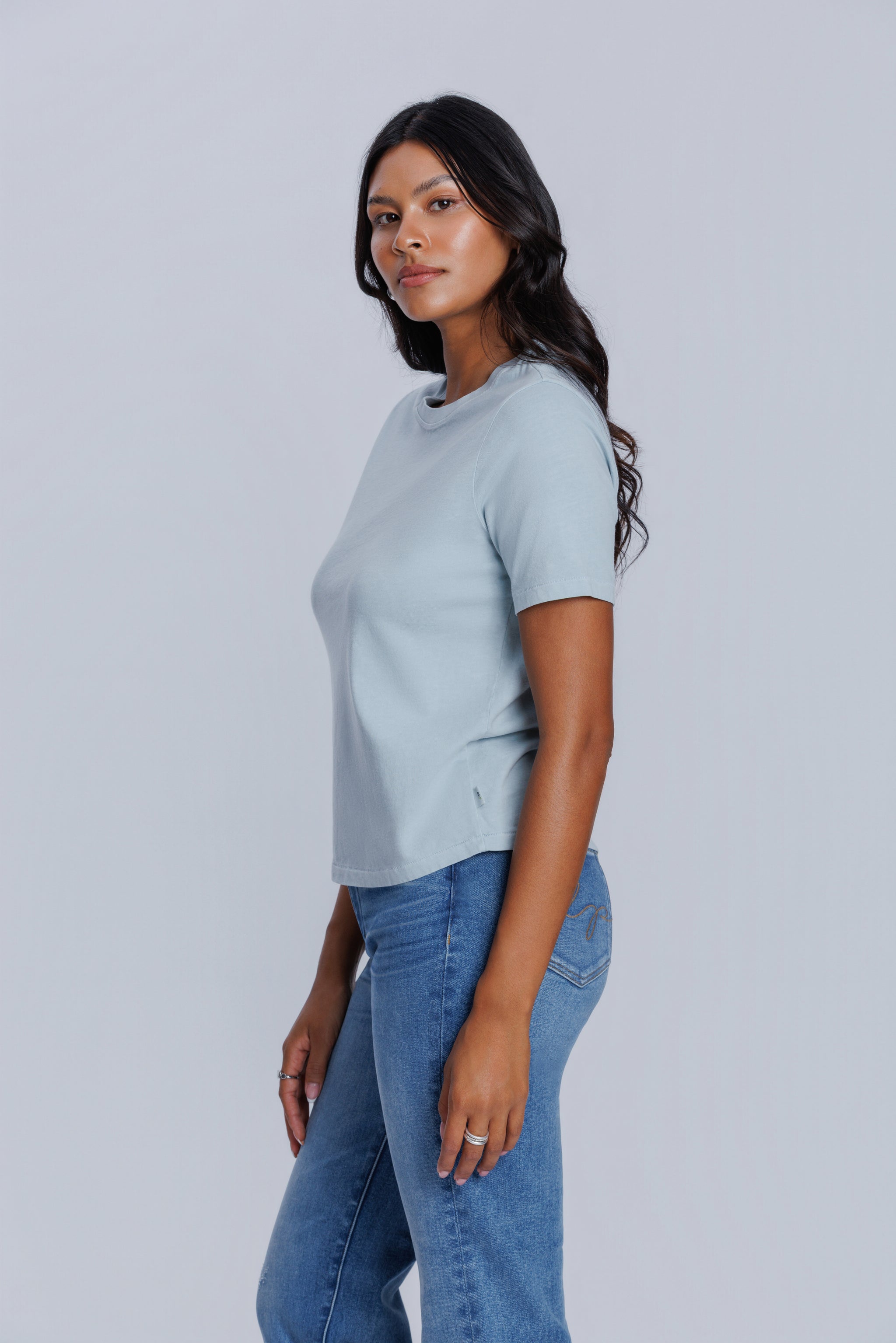 Woman wearing a DePart light blue t-shirt and blue jeans against a light gray background