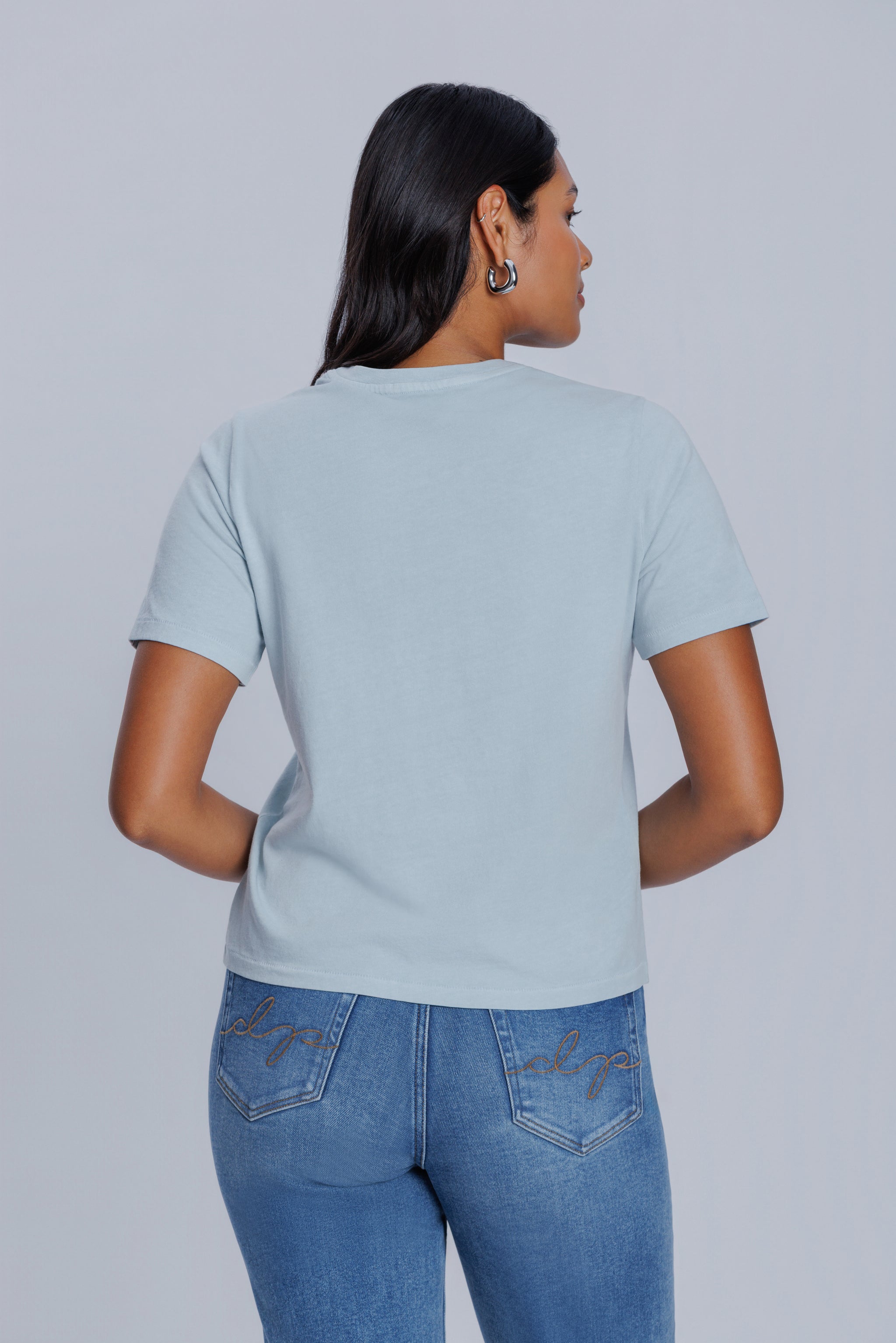 Woman wearing a DePart light blue t-shirt and blue jeans against a light gray background