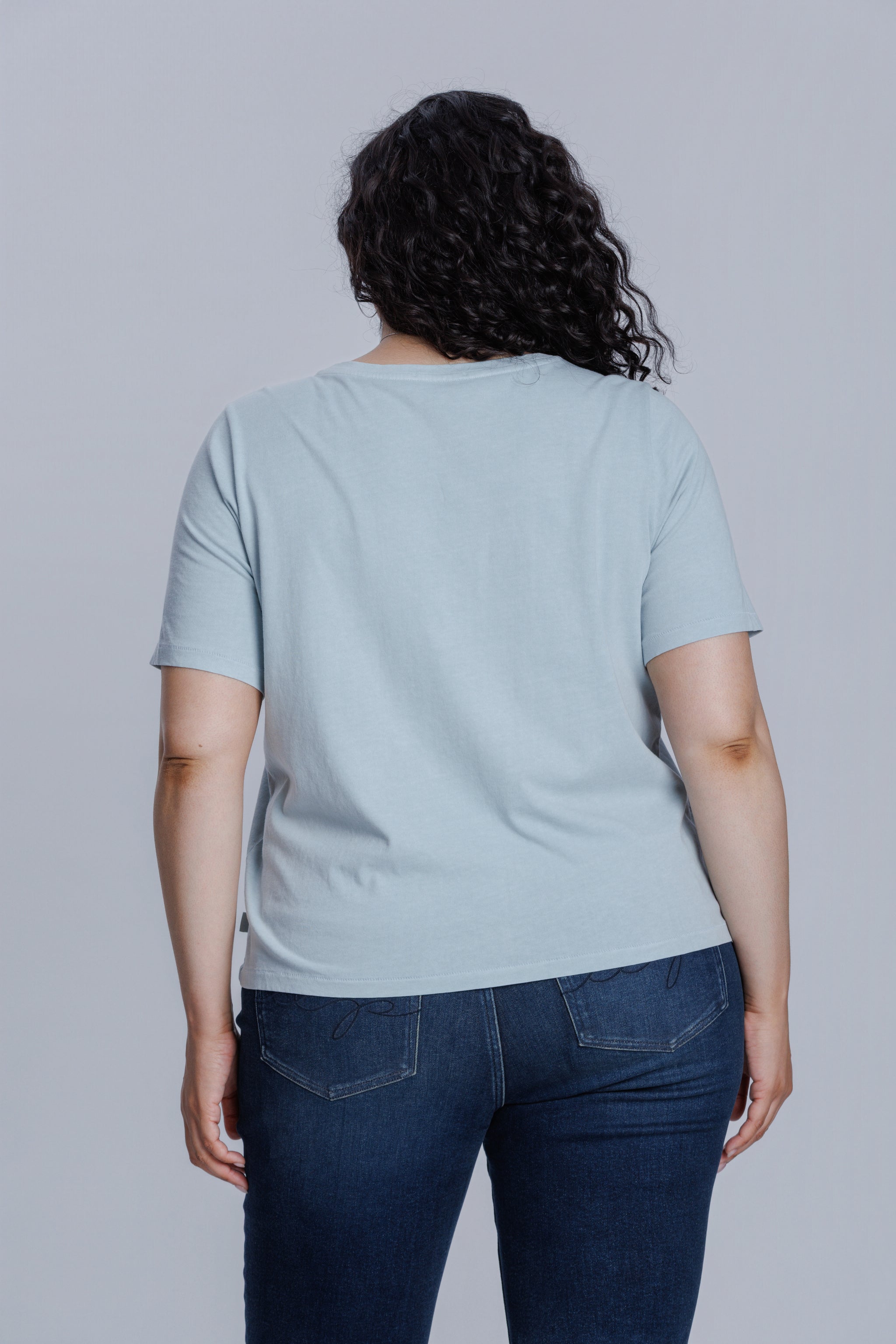 Woman wearing a DePart light blue t-shirt and dark blue straight jeans against a light gray background