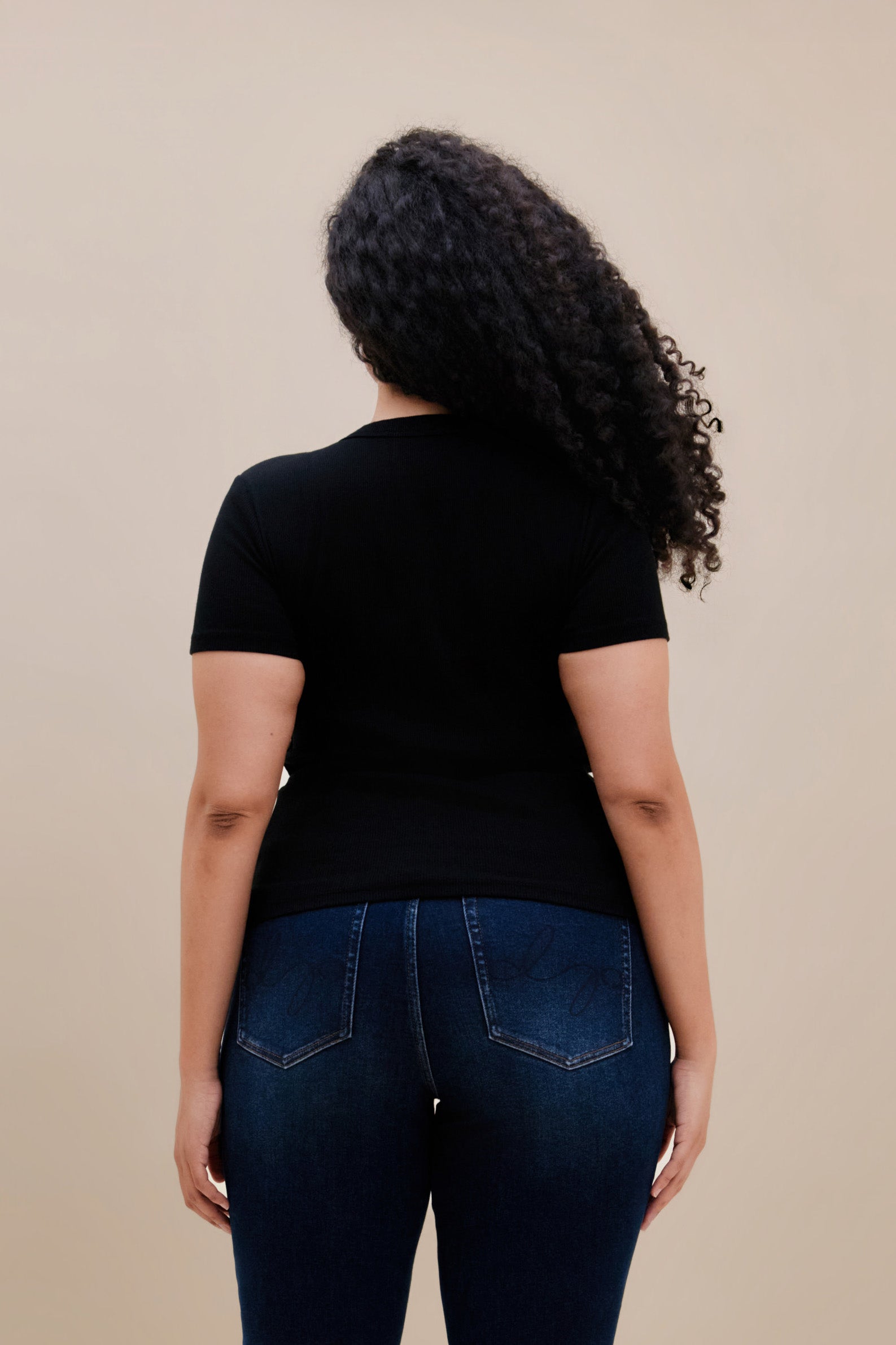 Backside of a woman wearing a fitted black rib tee and dark jeans against a beige wall