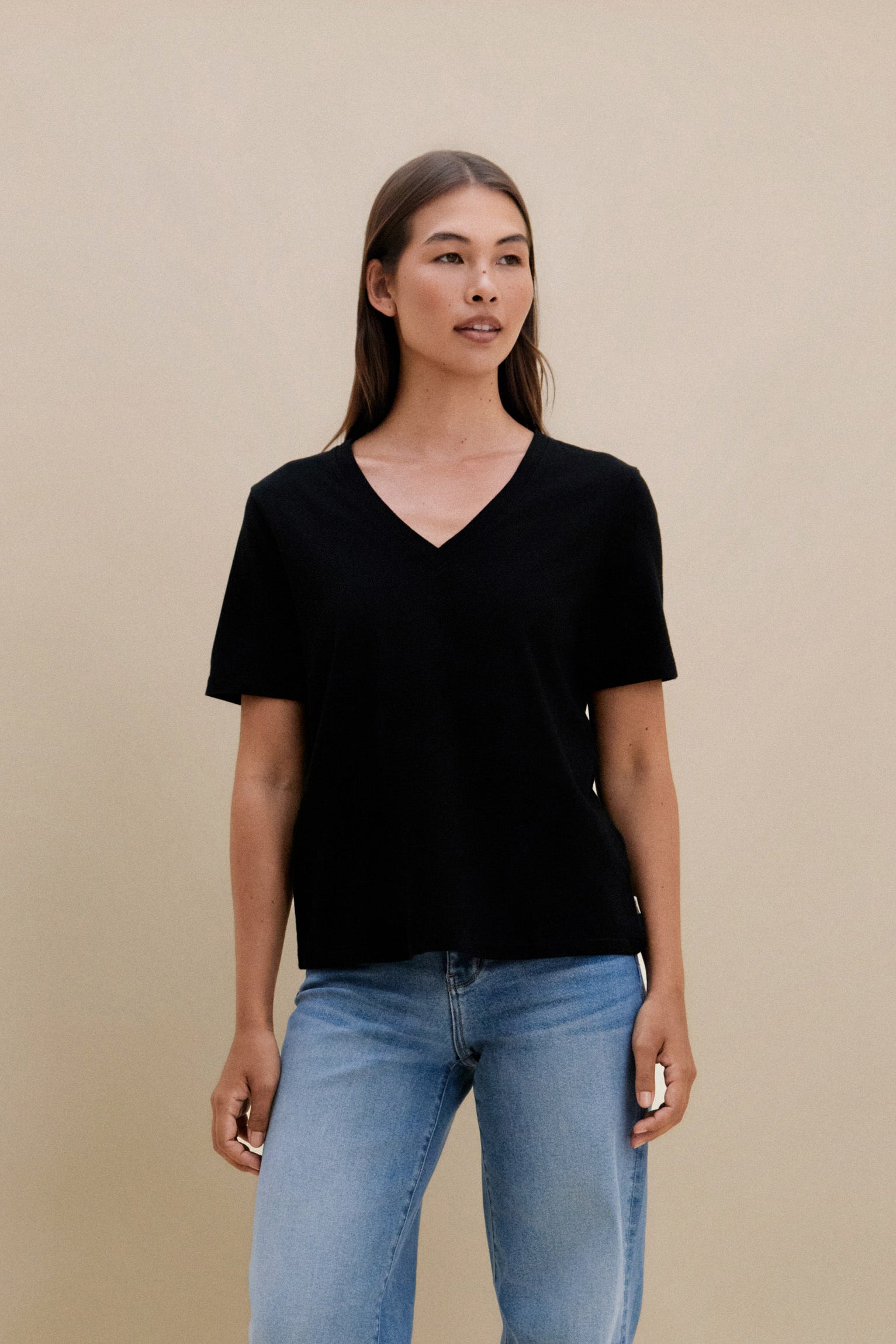 Woman wearing a black V-neck t-shirt and blue jeans against a beige background | model-size: 4