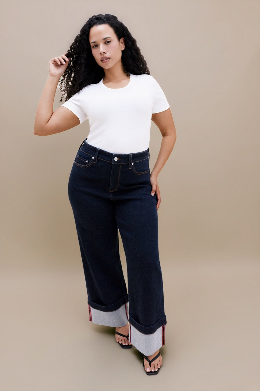 A woman modeling dark wash wide-leg cuffed jeans with a white t-shirt, standing against a solid beige studio background.
