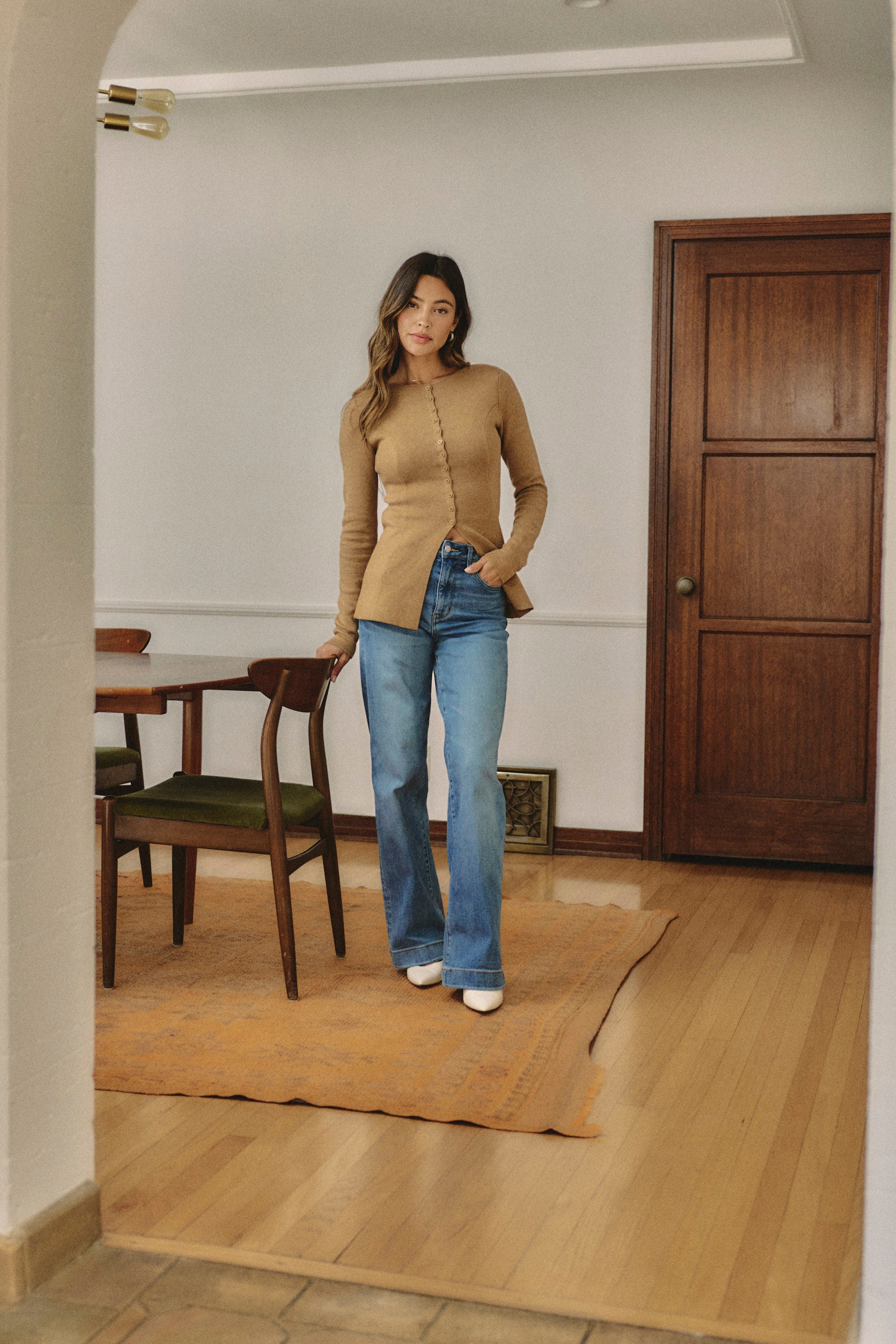 Woman standing in a room with wooden floor and furniture wearing a camel colors top and jeans | model-size: 4