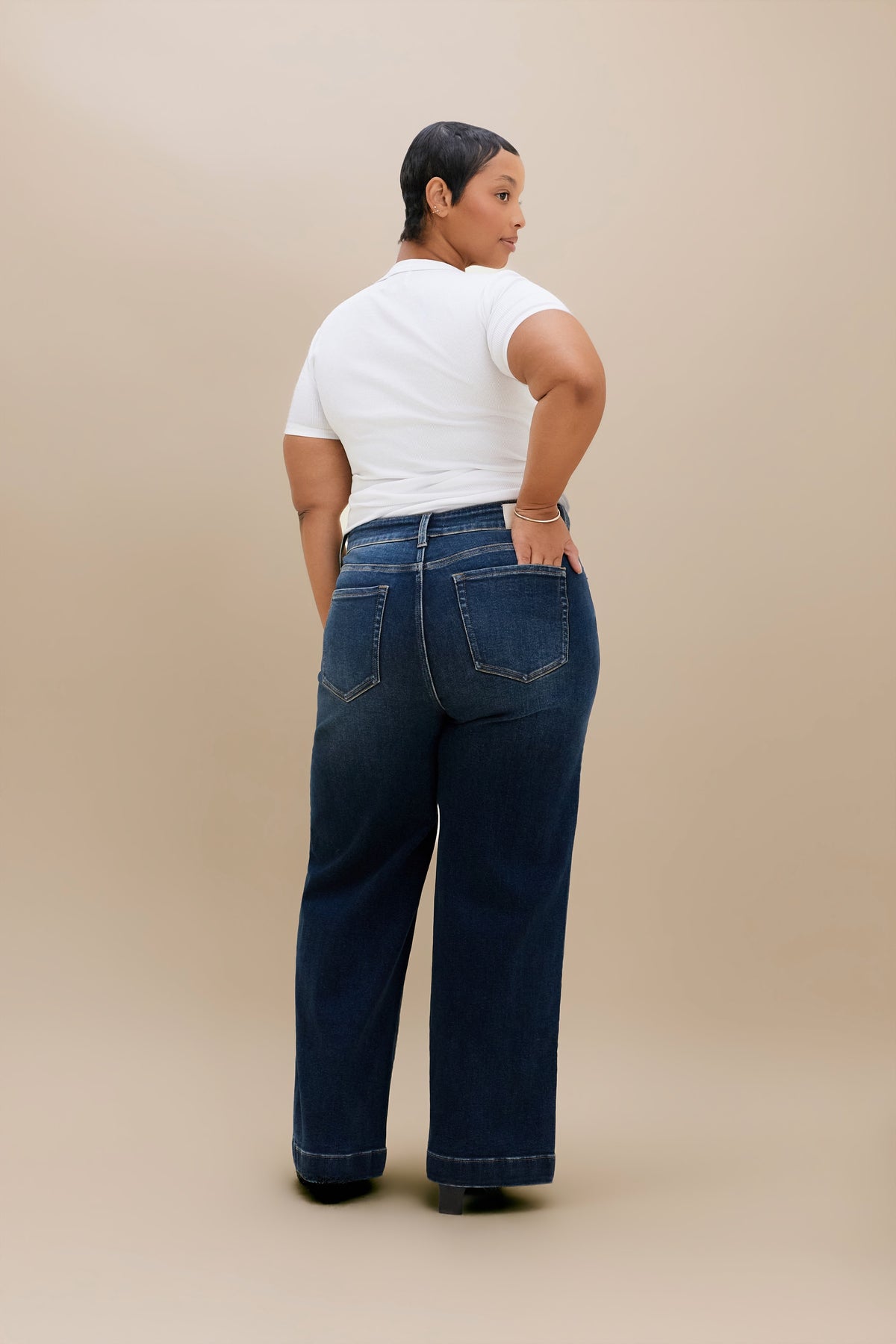 A model shown from the back wearing dark blue wide-leg jeans and a white t-shirt, standing against a solid beige studio background.