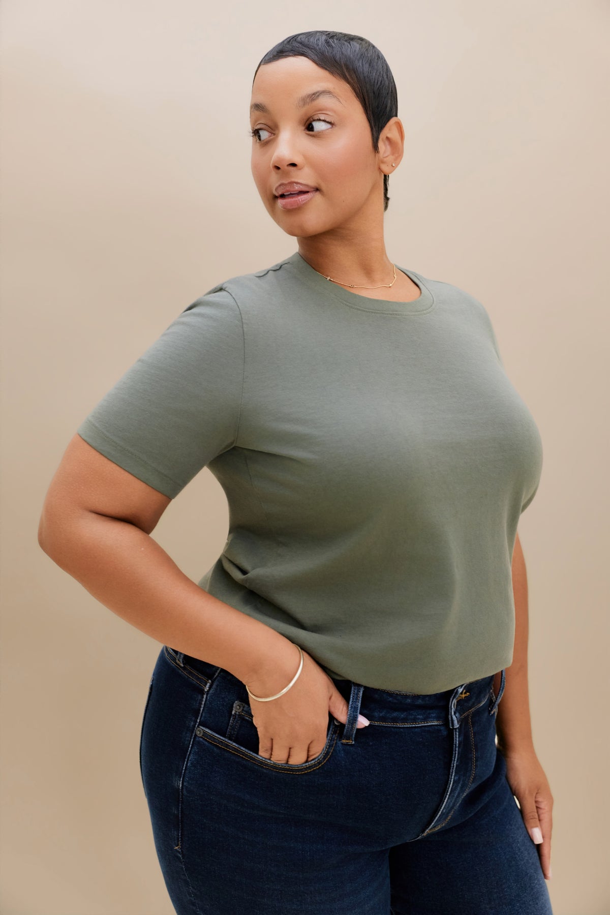 A woman modeling a short-sleeve crewneck t-shirt in sage green, paired with dark blue jeans, against a solid beige studio background.
