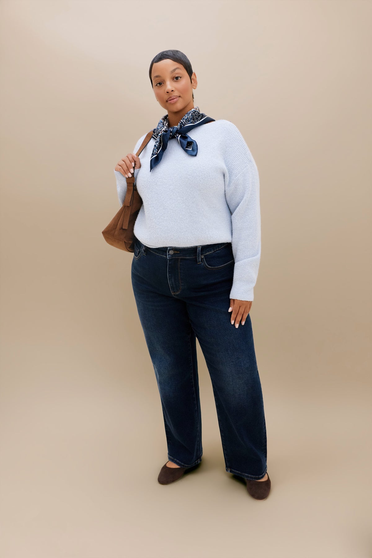 A woman modeling a light blue knit sweater, dark blue straight leg jeans, a navy and white patterned neck scarf, and brown flats, holding a brown shoulder bag against a solid beige studio background.