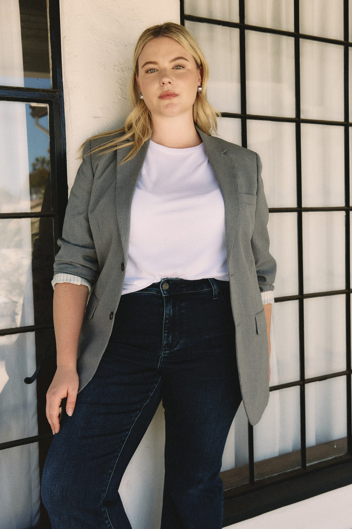 Woman wearing a gray blazer over a white shirt and dark jeans, standing against a windowed wall | model-size: 16