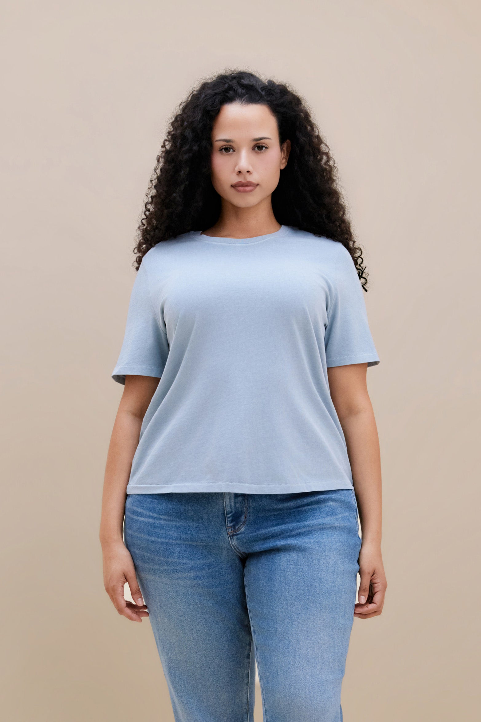 Woman wearing a blue crew neck tee and jeans with a beige background | model-size: 16