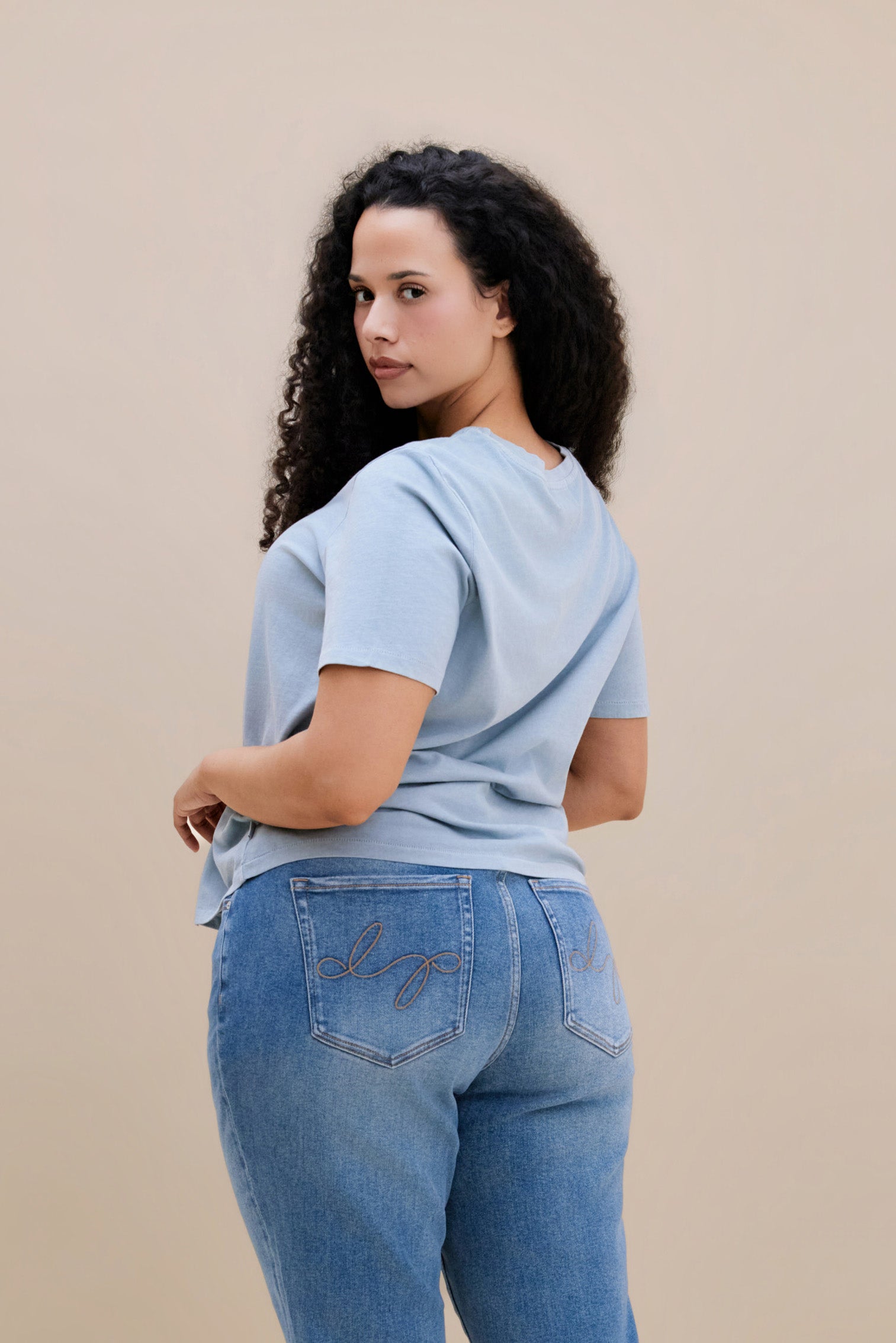 Backside of a woman wearing a blue crew neck tee and jeans with a beige background