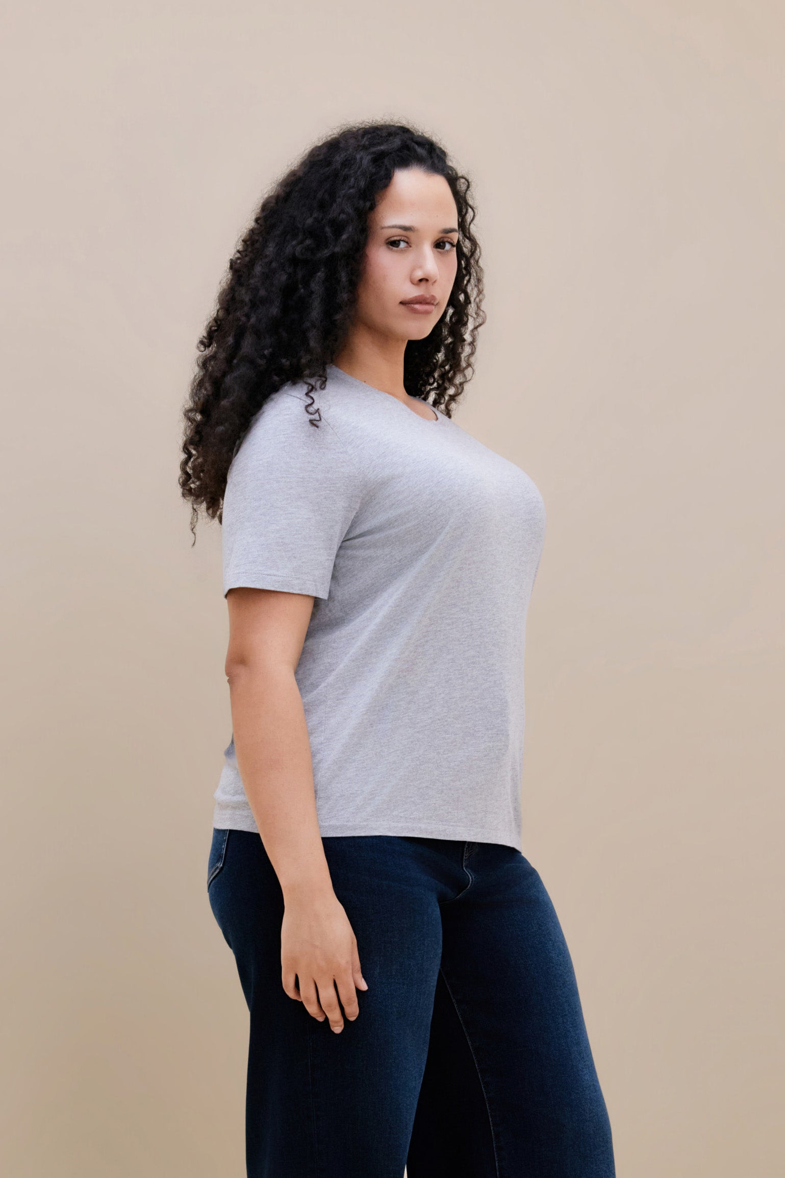 Woman wearing a light heather grey t-shirt & dark jeans against a beige background