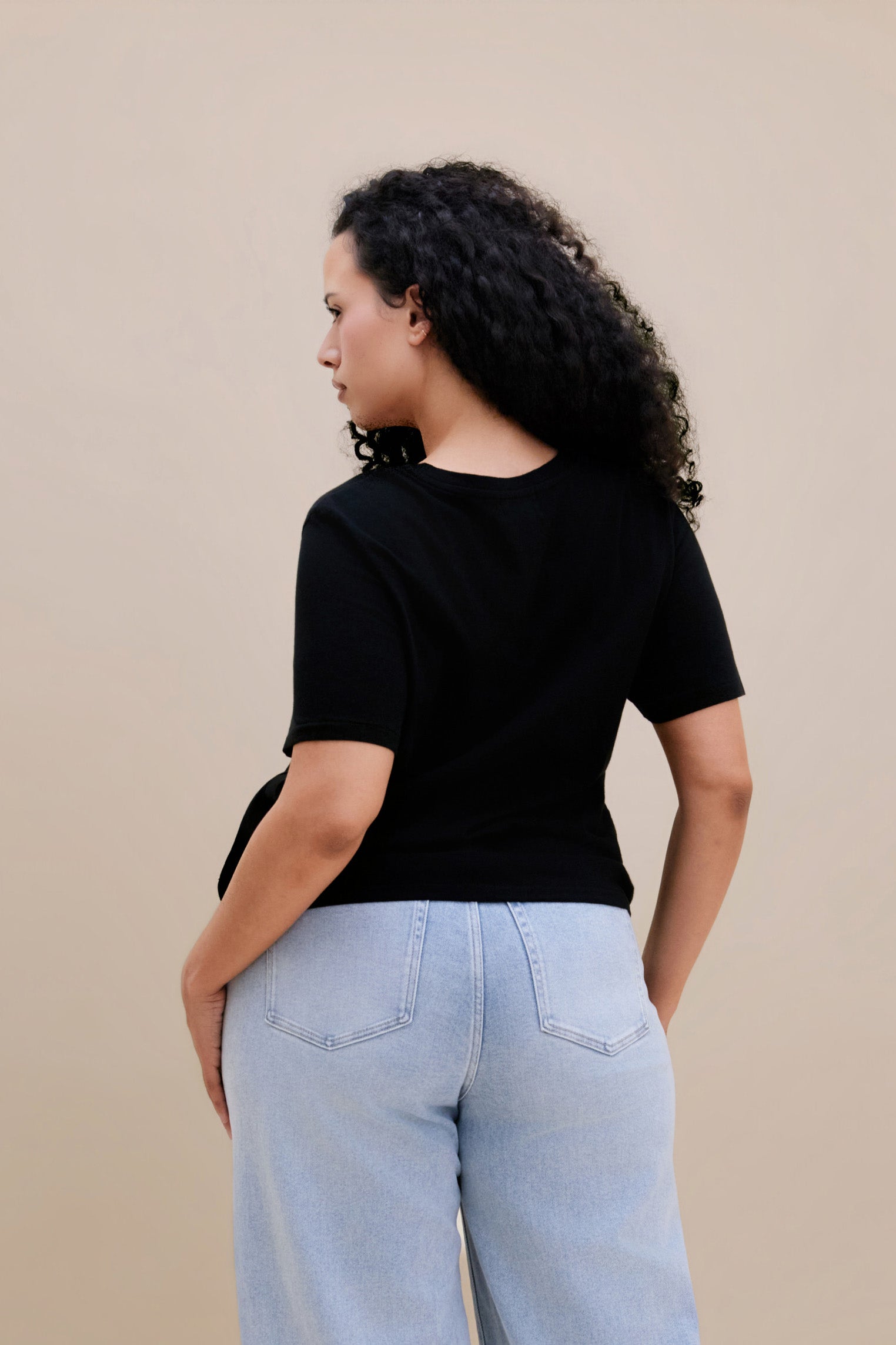 Backside of a woman wearing a black t-shirt and light blue jeans against a beige background