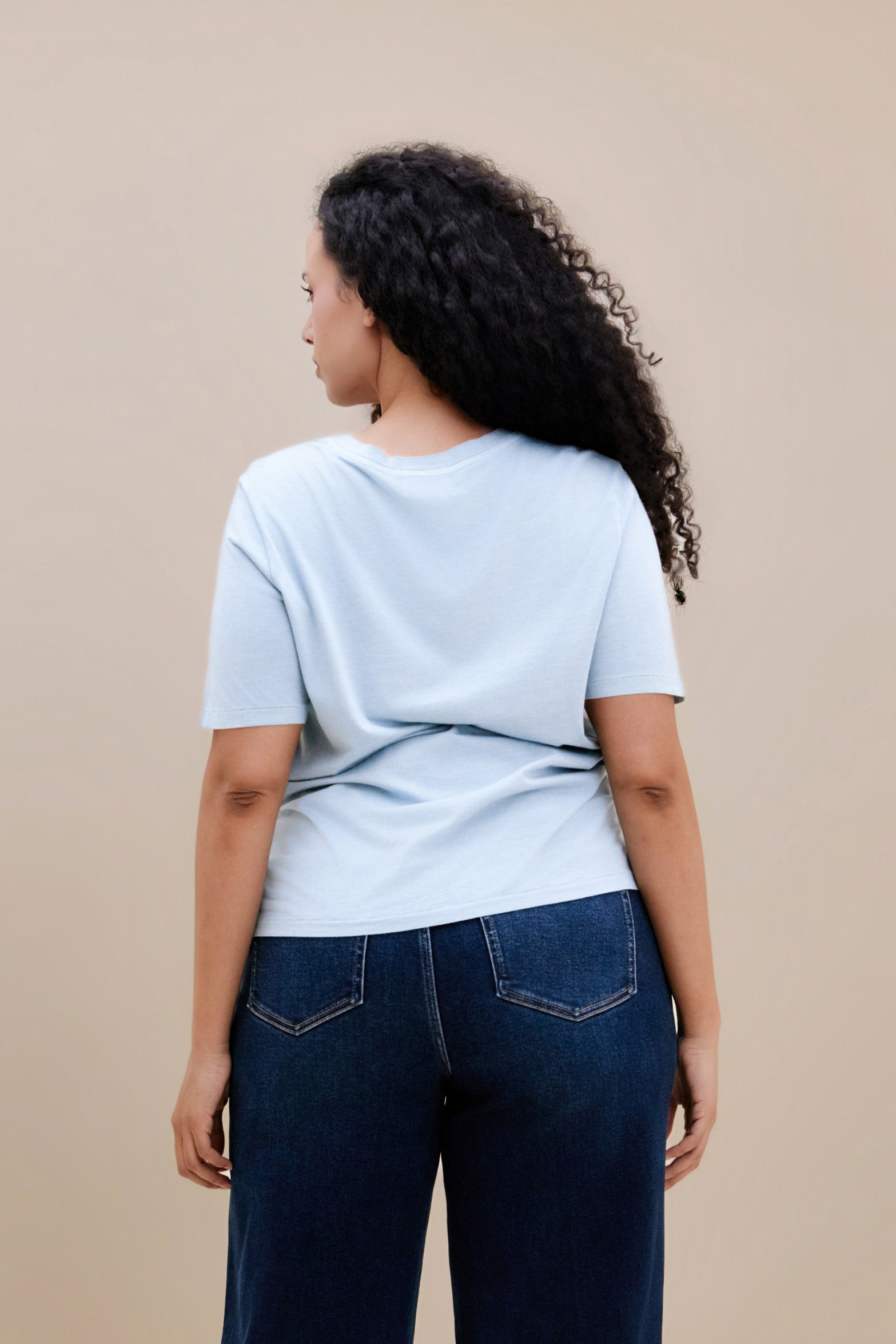 Backside of a woman wearing a light blue t-shirt and dark jeans against a beige background