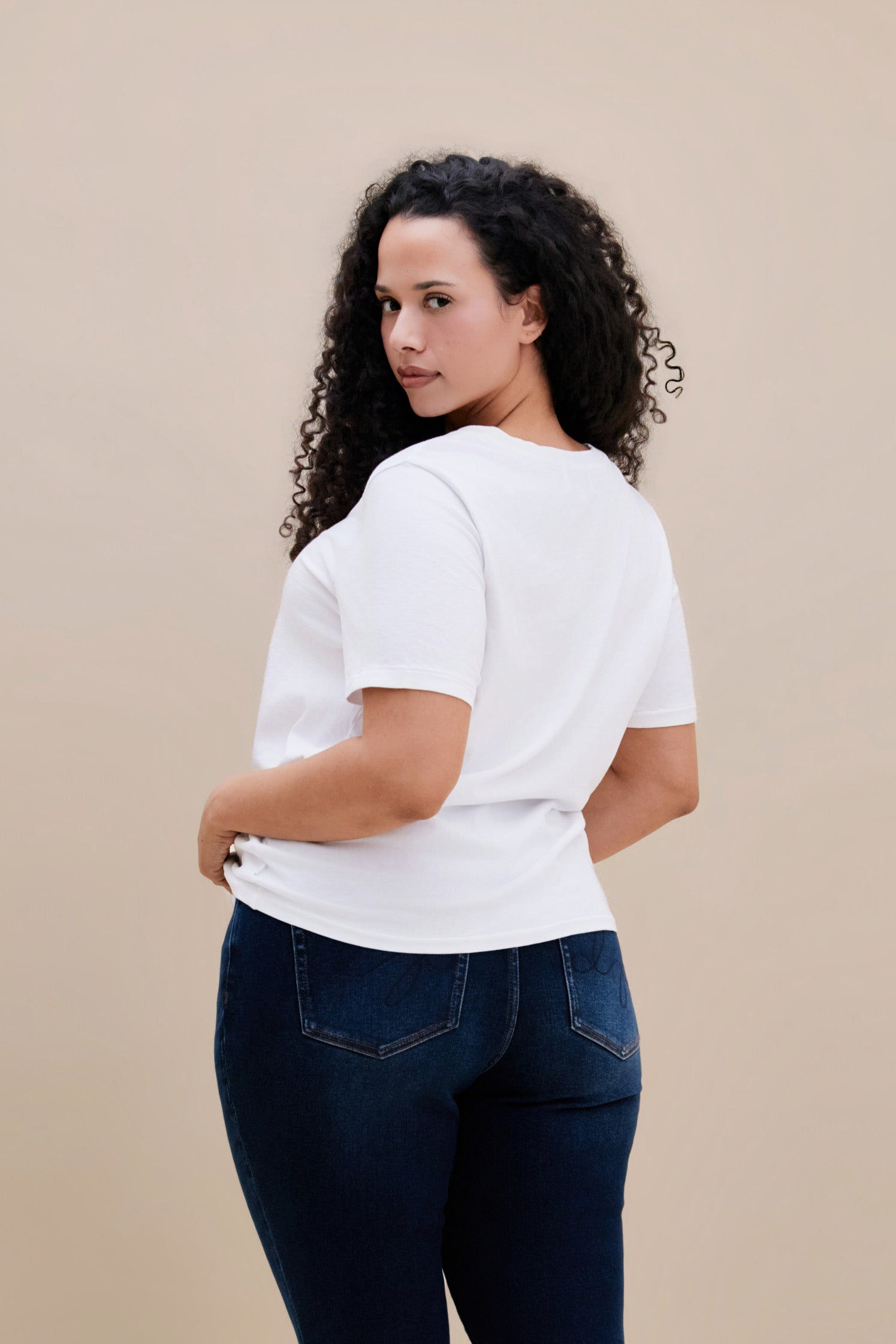 Backside of a woman wearing a plain white t-shirt and dark jeans against a beige background