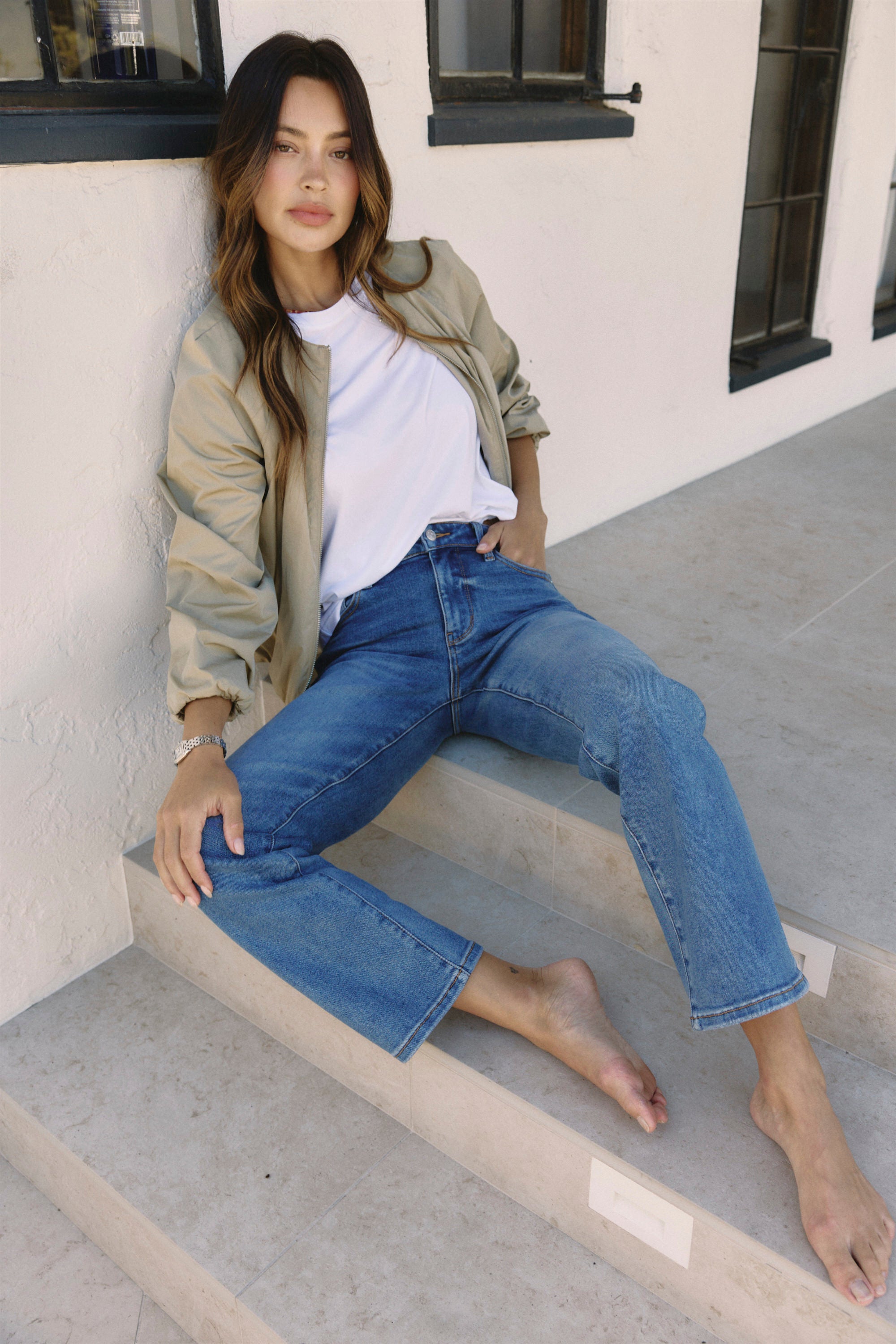 Woman sitting on steps wearing a beige jacket, white shirt, and blue jeans | model-size: 4