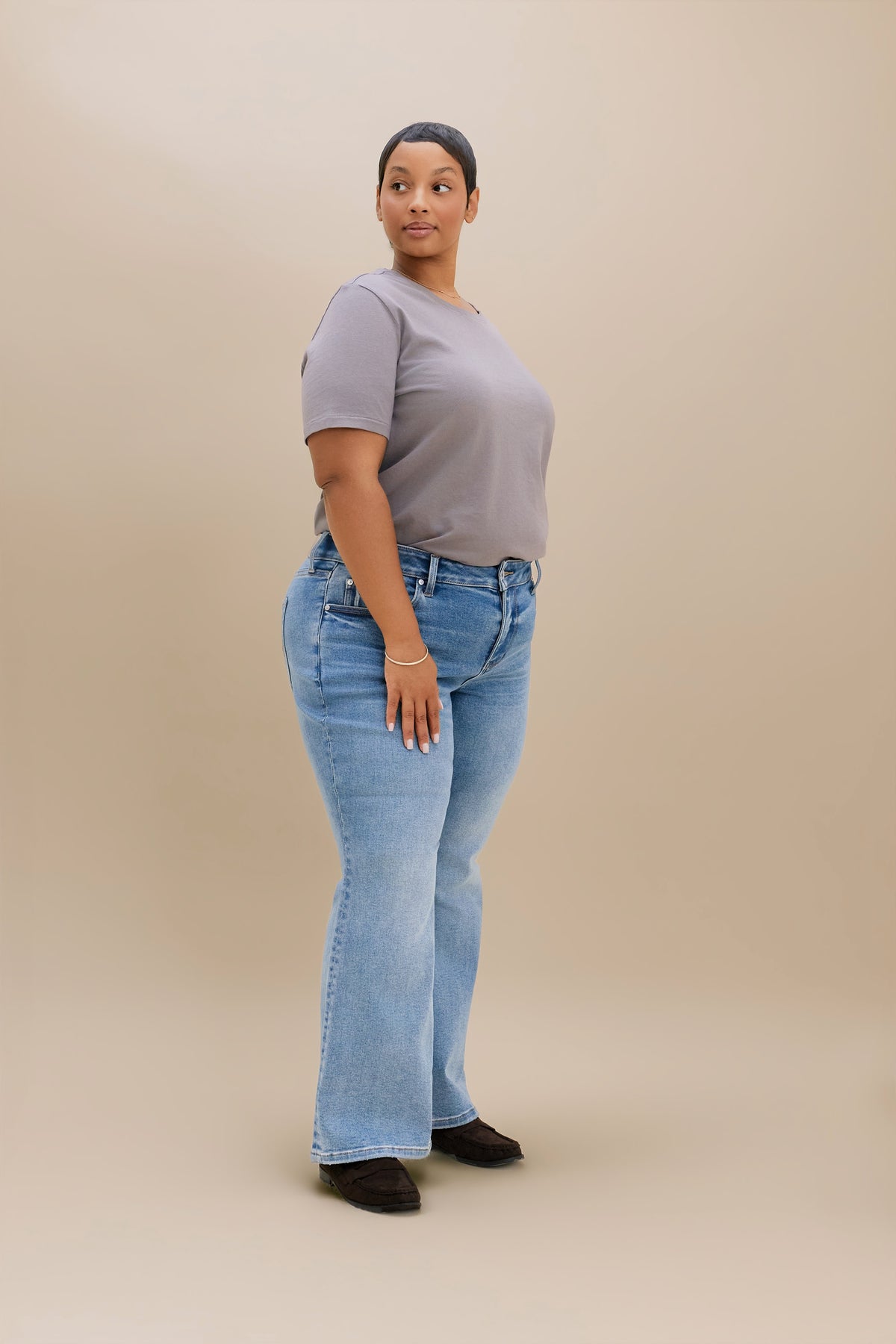 A woman modeling light-wash blue boot cut jeans with a gray t-shirt, standing against a solid beige studio background.