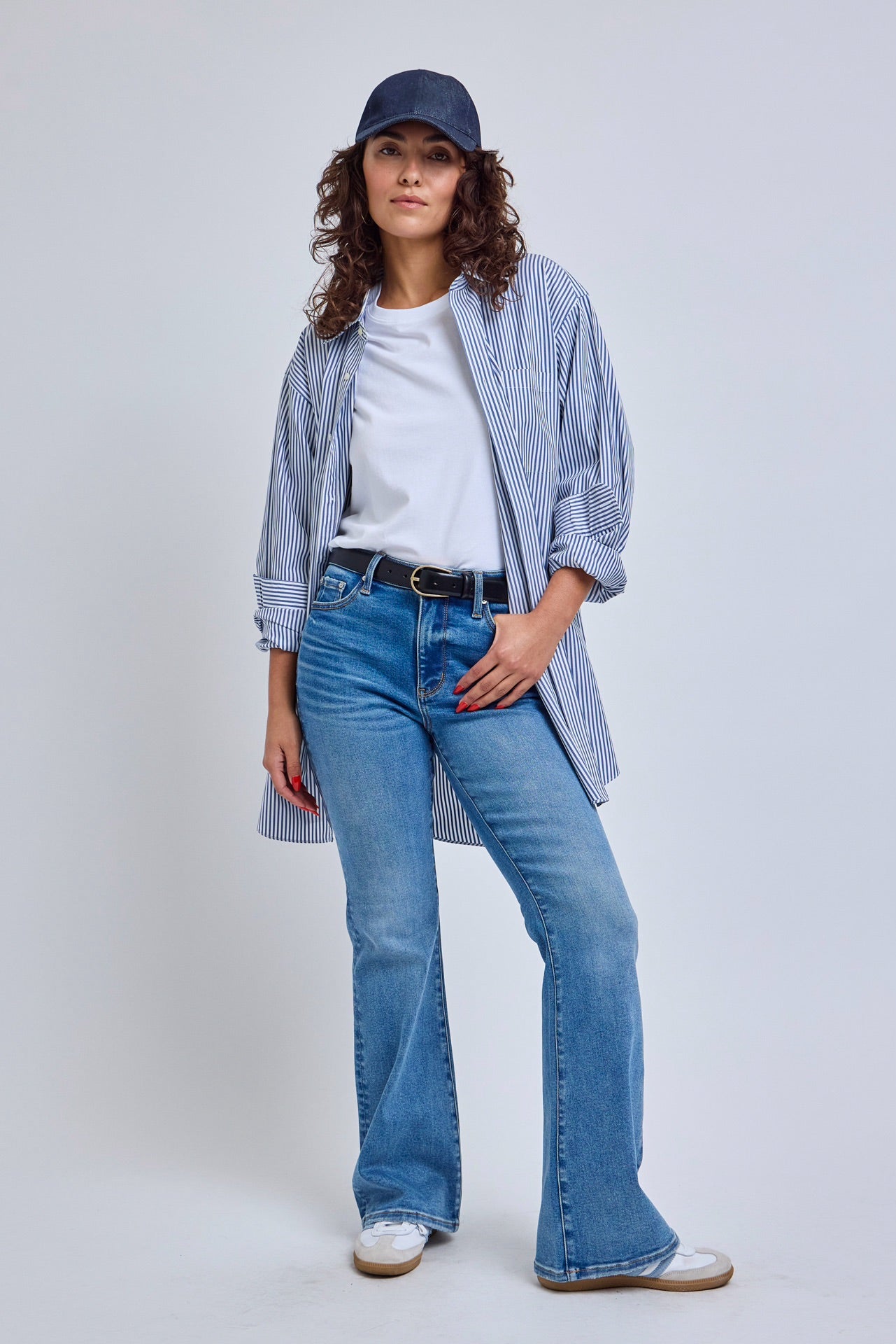 Woman wearing a blue and white striped shirt, white sneakers, and a navy cap on a light gray background | model-size: 4