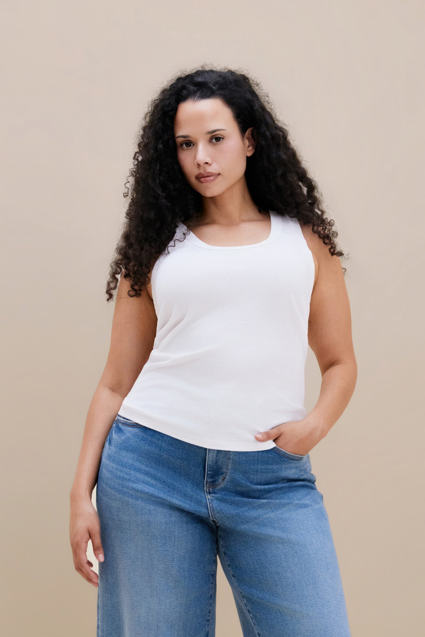 Woman wearing a white ribbed tank top and jeans against a beige background | model-size 16