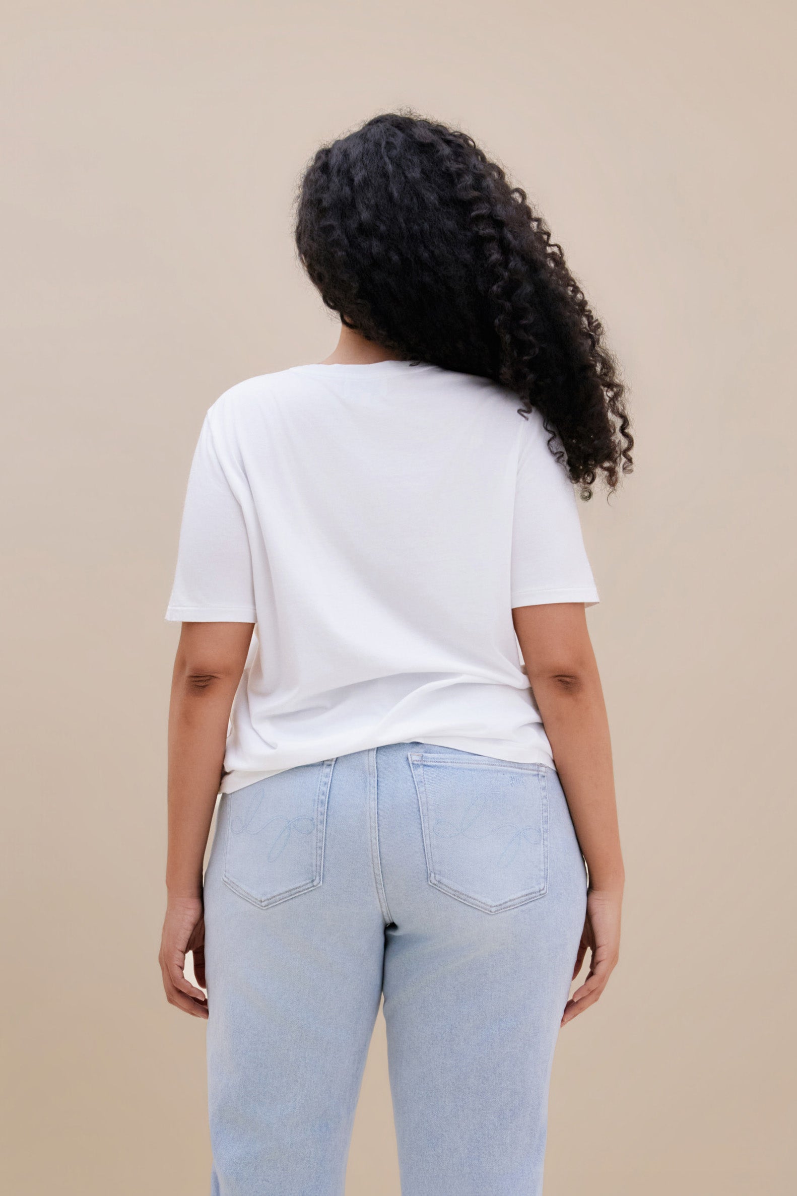 Backside of a woman wearing a white t-shirt and light blue jeans against a beige background