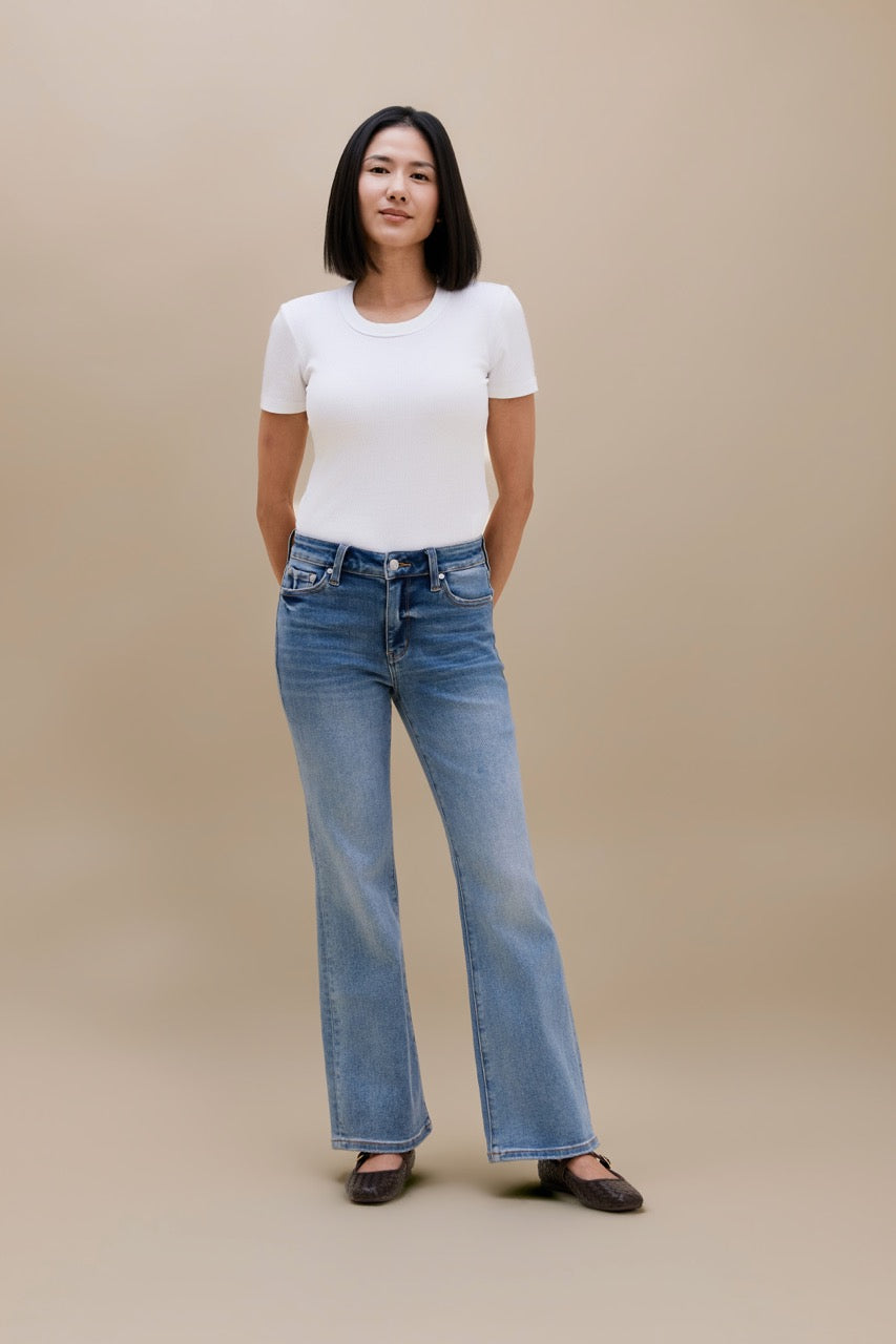 A woman modeling a white short-sleeve crewneck t-shirt and light-wash blue flare jeans, standing against a solid beige studio background.