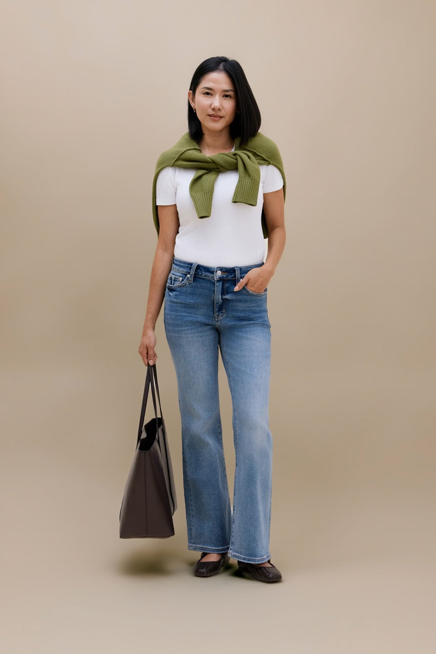 A woman stands against a plain beige studio background wearing a white short-sleeve top, blue high-waisted flare jeans, and dark flats. She has an olive green knit wrap tied around her shoulders and is holding a dark taupe tote bag.