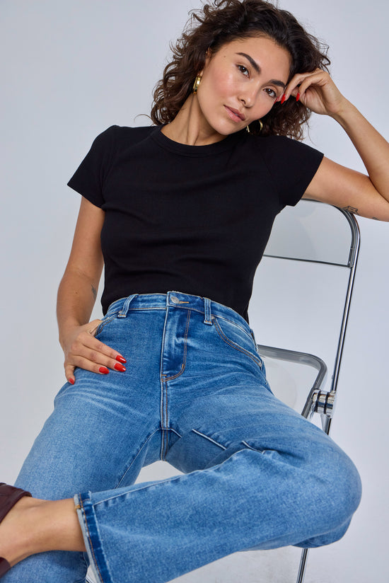Woman wearing a black top and blue jeans sitting on a chair against a plain background | model-size: 4