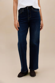 A person wearing dark blue straight-leg jeans, a white top, and dark loafers, standing against a plain beige studio background.