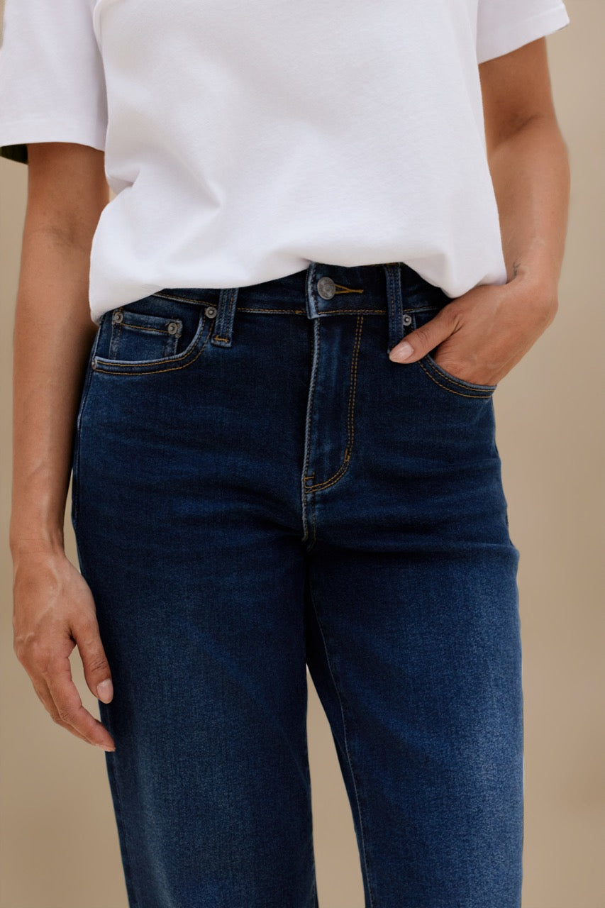 A person wearing dark blue denim jeans and a white short-sleeve top, shown from the chest down against a plain beige background.