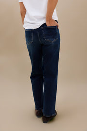 A person shown from the back wearing dark blue straight-leg jeans, a white t-shirt, and brown shoes against a solid beige studio background.