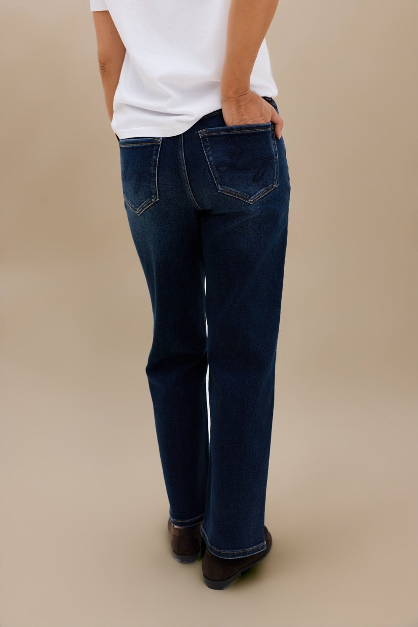 A person shown from the back wearing dark blue straight-leg jeans, a white t-shirt, and brown shoes against a solid beige studio background.